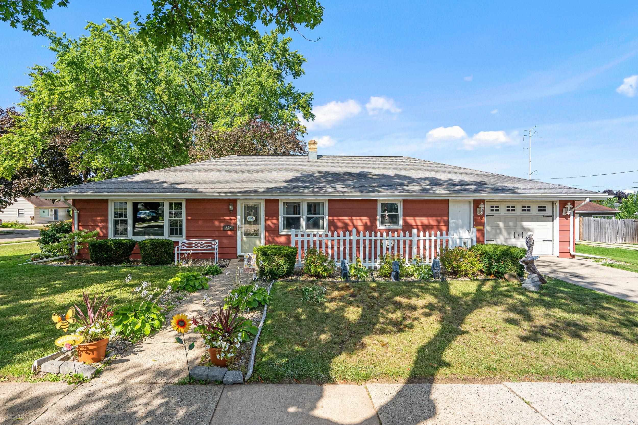 Red single-story home with attached garage, white picket fence, and flower-lined front walkway.