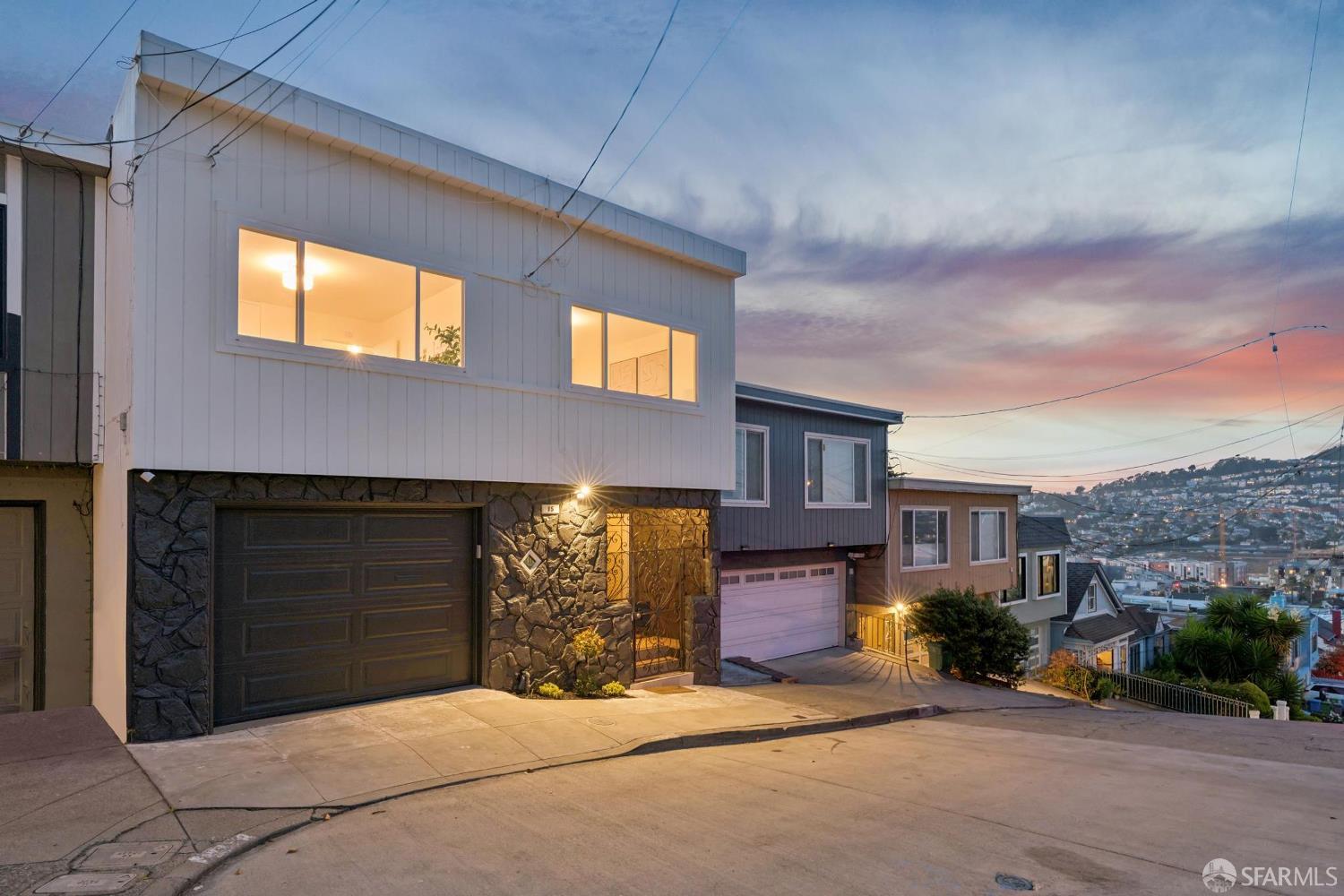 Modern two-story home with a stone facade and glowing windows on a sloped city street at dusk.