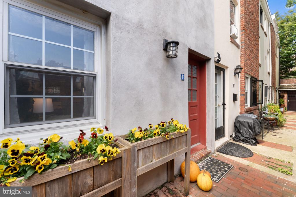 Townhouse entrance with a red door, flower-filled wooden planters, and pumpkins beside a brick walkway.