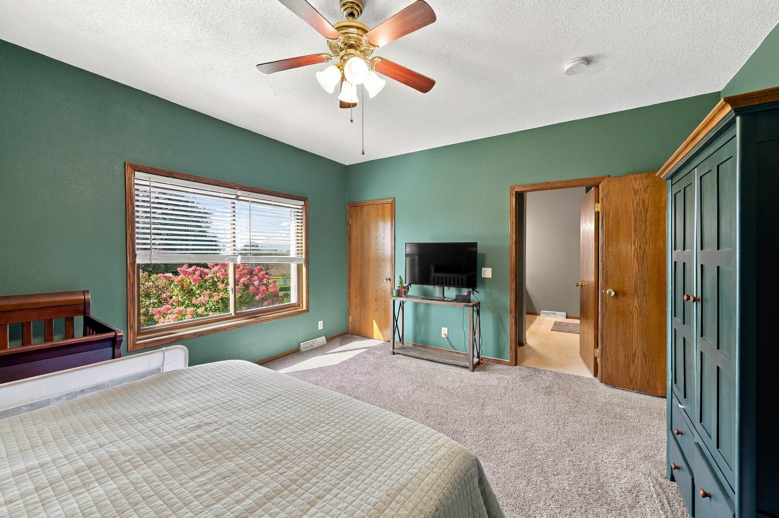 Bedroom with teal walls, a large window overlooking flowering shrubs, and a ceiling fan.