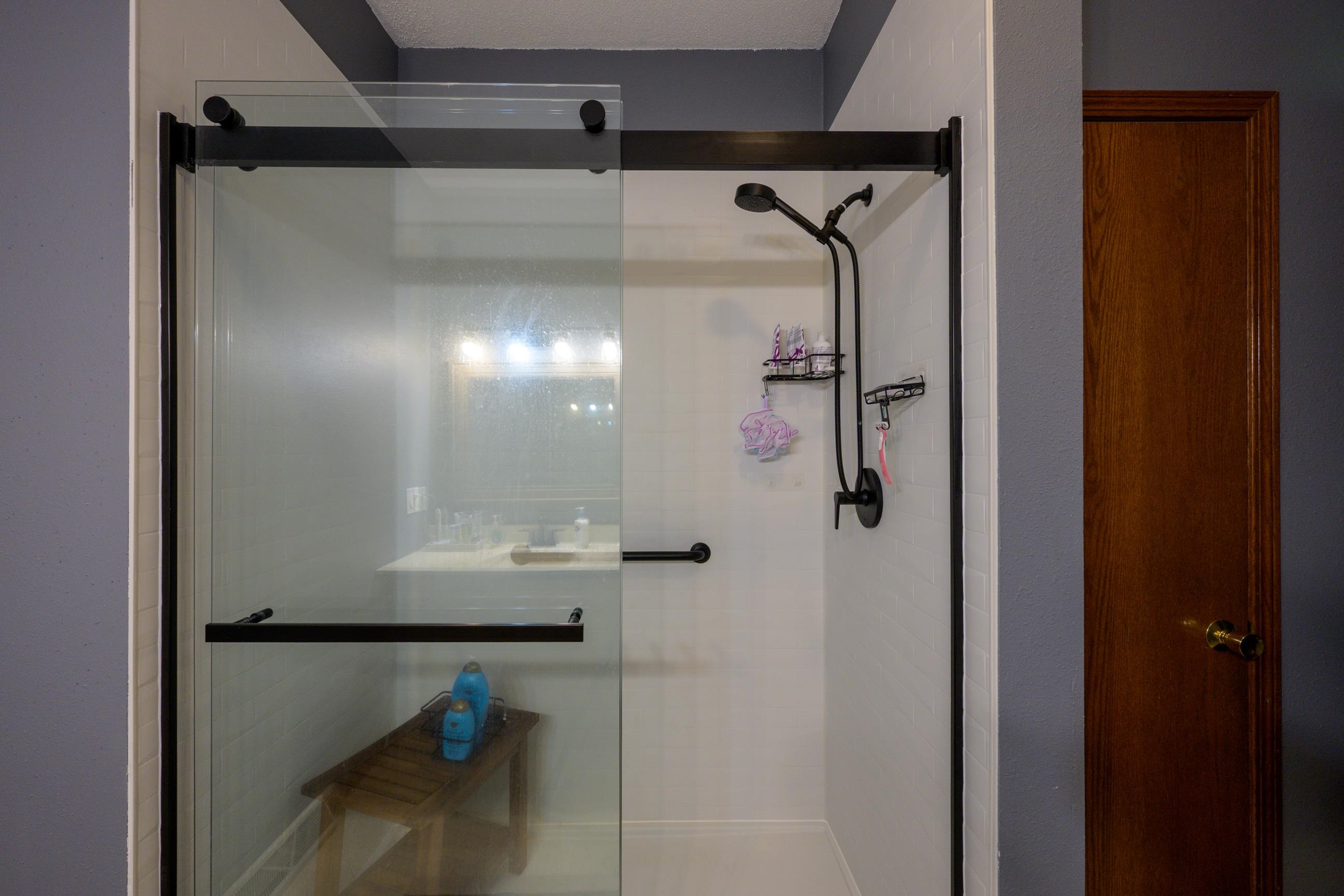 Walk-in shower with a black-framed glass enclosure, white tile, and handheld showerhead.