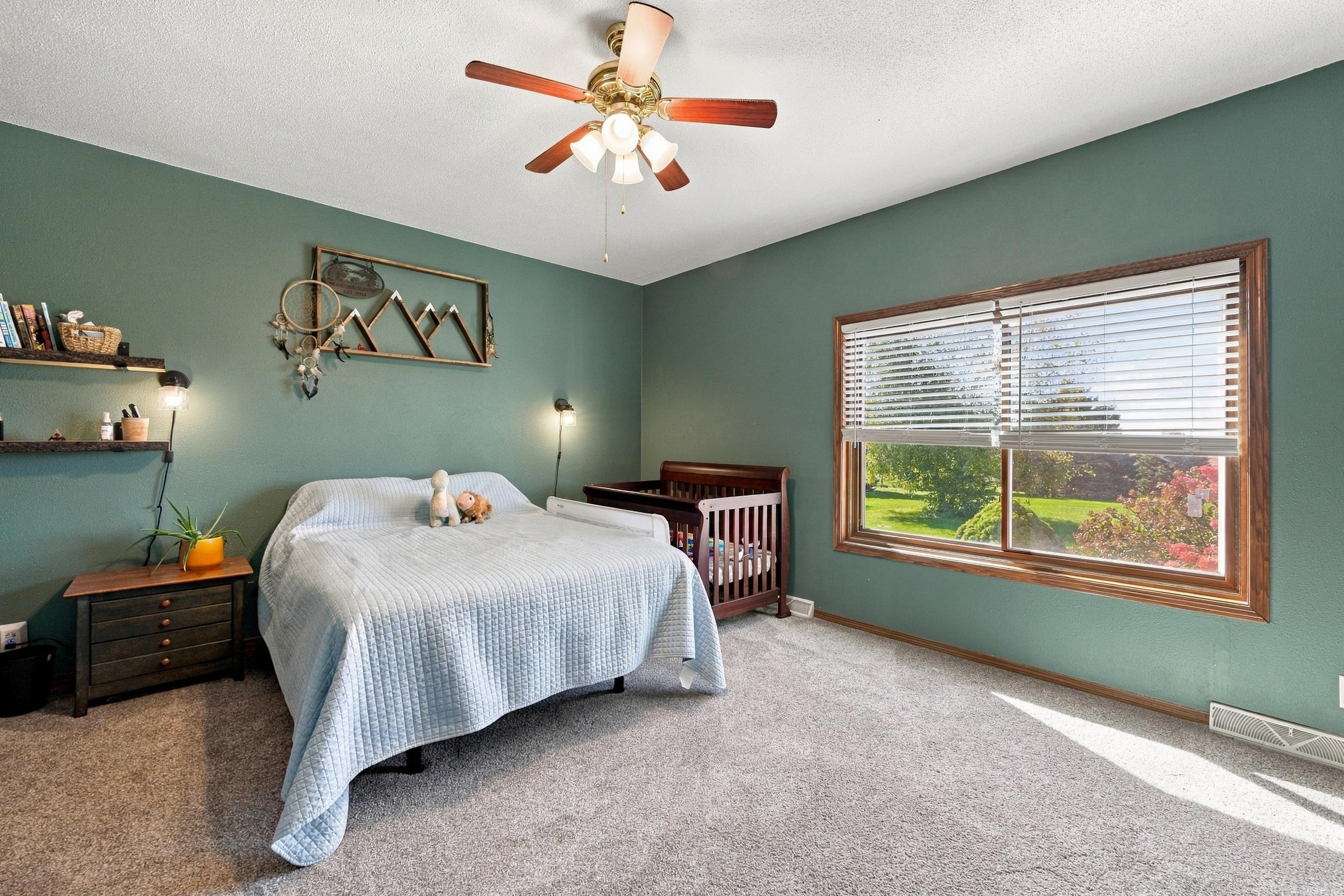 Bedroom with a bed, wooden crib, teal walls, and a large sunlit window overlooking the yard.