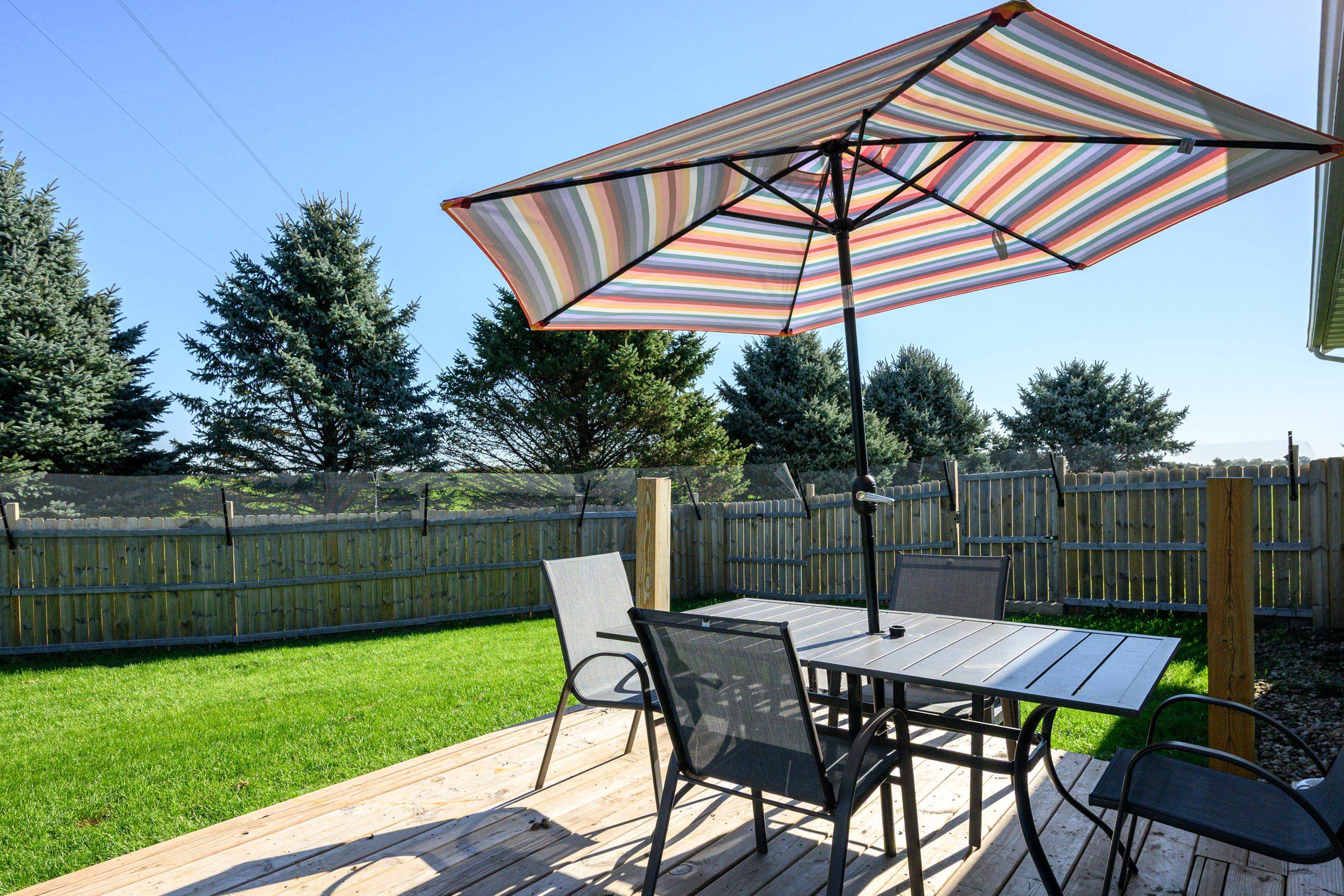 Backyard deck with outdoor dining set beneath a striped umbrella, overlooking a fenced lawn and evergreens.