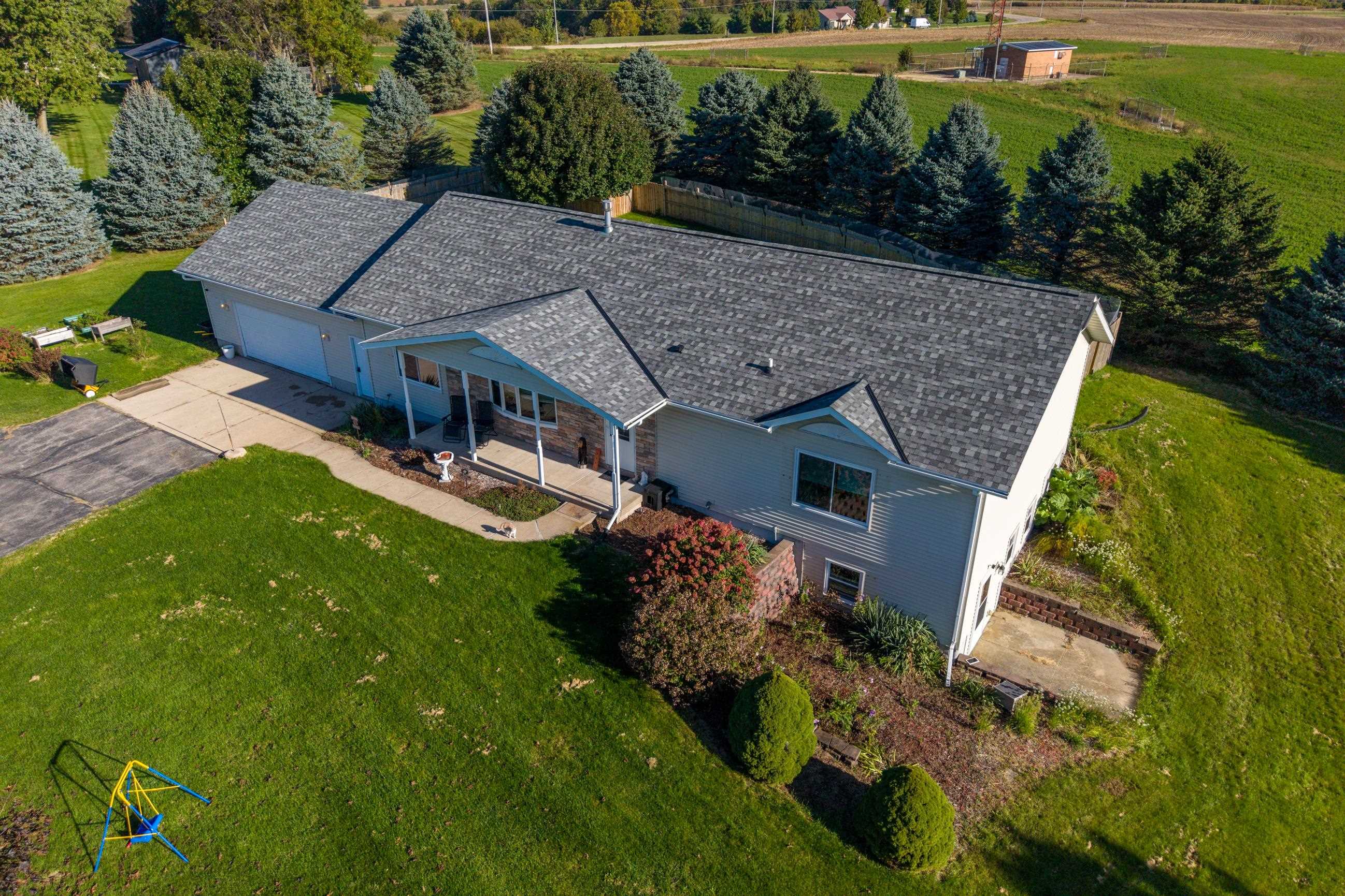 Aerial view of a gray ranch home with a covered patio, broad lawn, and mature evergreen trees.