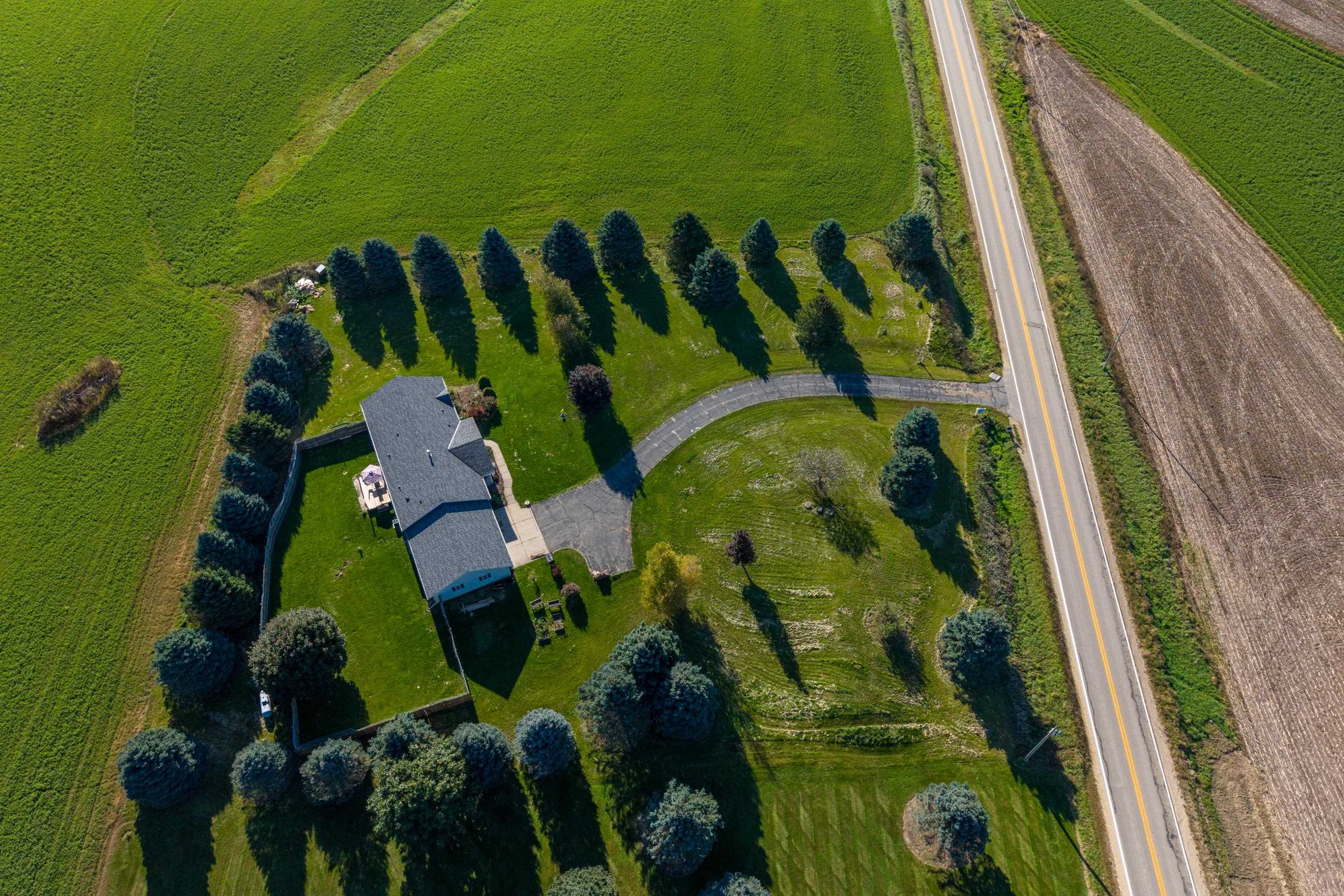 Aerial view of a home with a tree-lined lawn, curved driveway, and adjoining fields beside a highway.