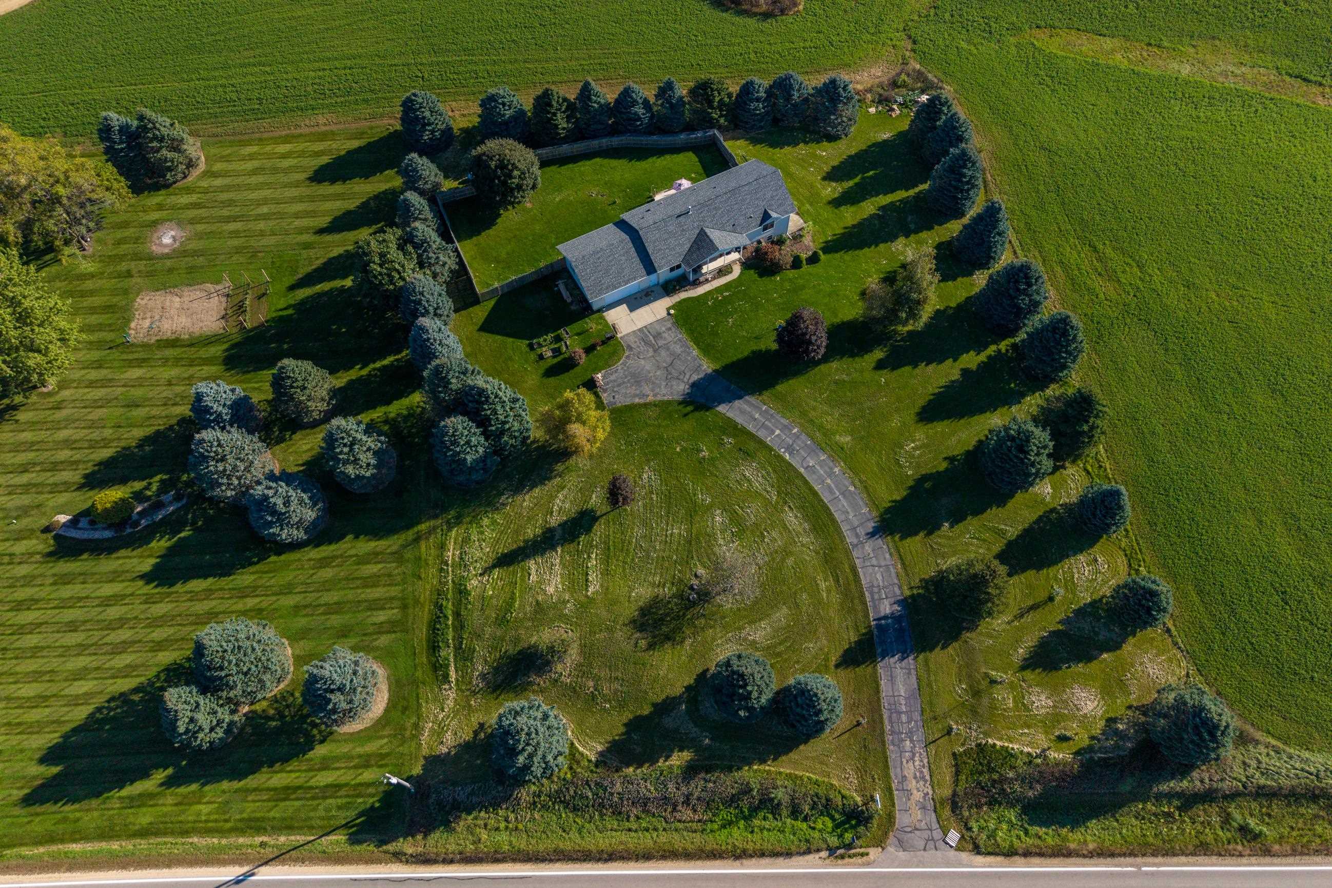 Aerial view of a home with a curved driveway, fenced backyard, and expansive tree-lined lawn.
