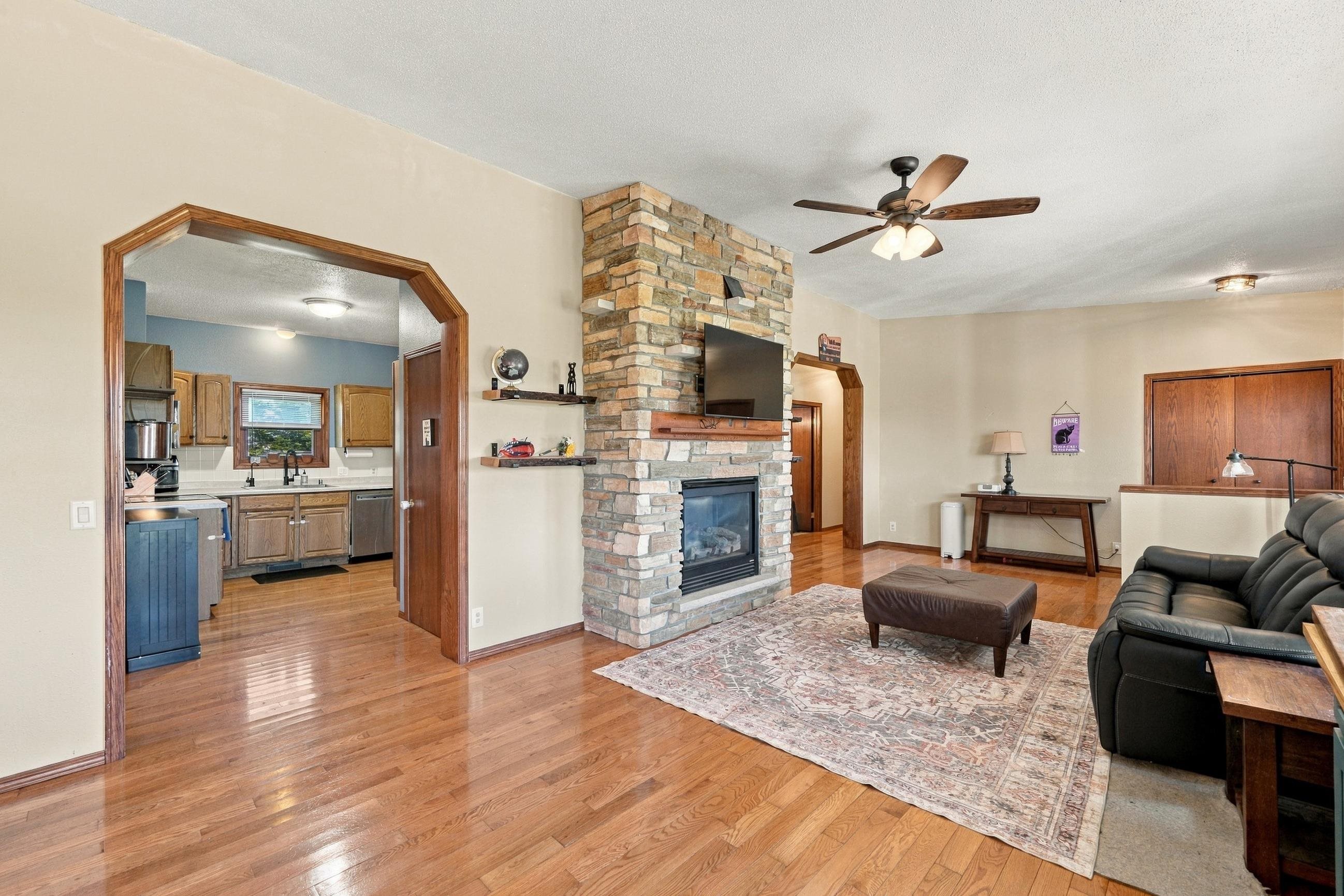 Living room with a stone fireplace, hardwood floors, and an open view into the kitchen.