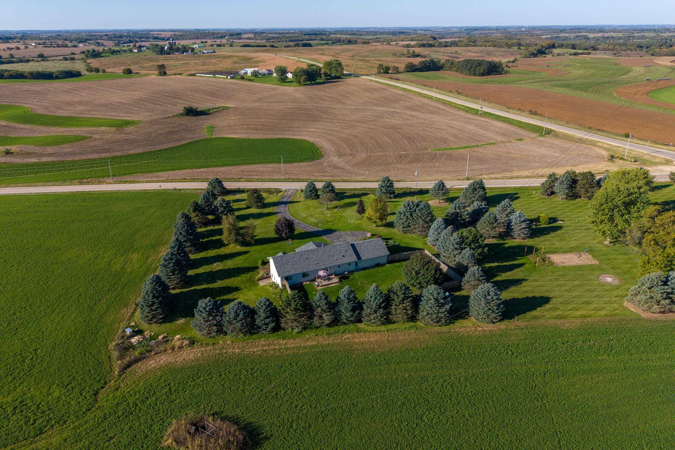 Rural home surrounded by evergreen trees and lawns, with expansive farmland and a road beyond.