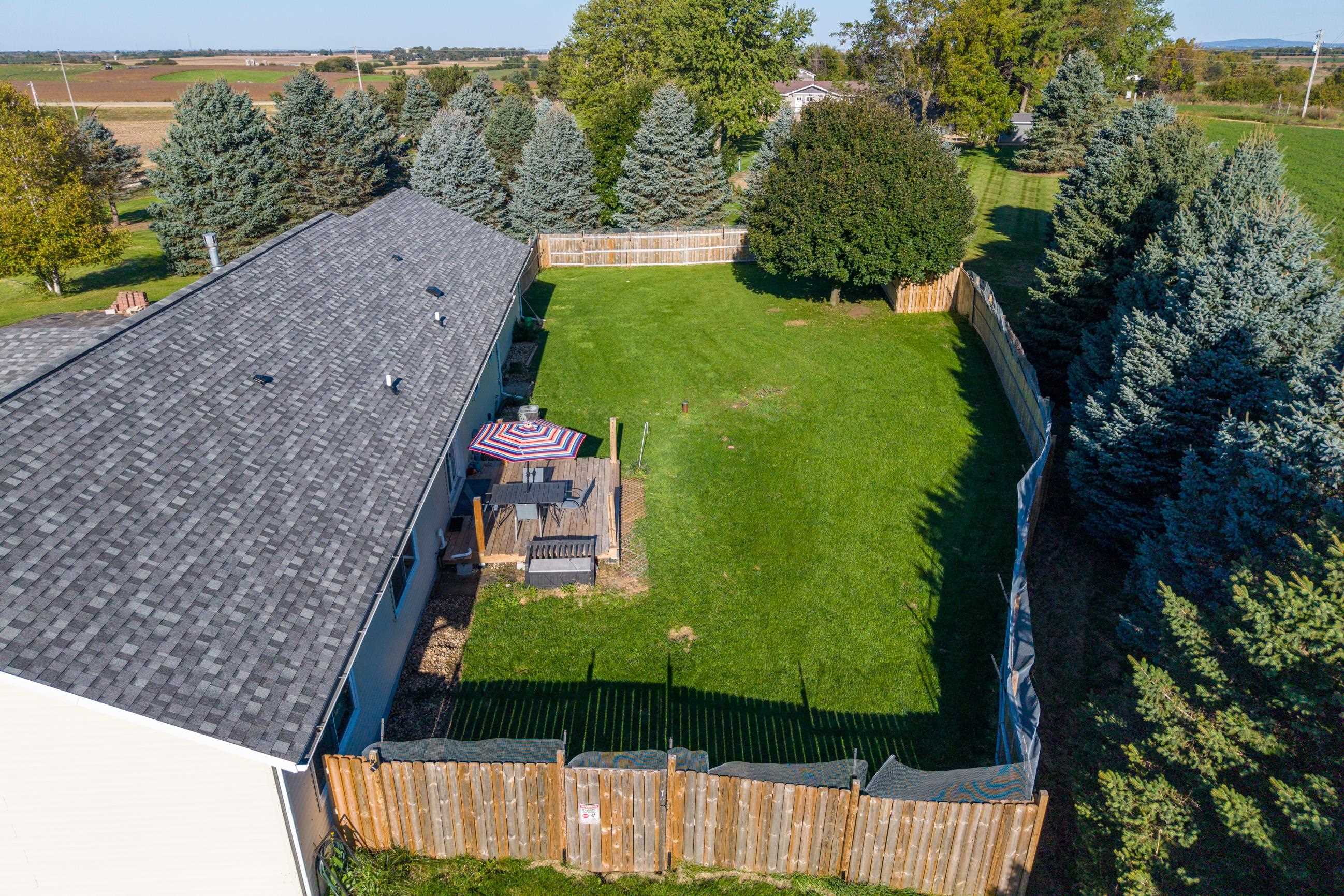 Aerial view of a fenced backyard with a deck, striped umbrella, and mature evergreen trees.