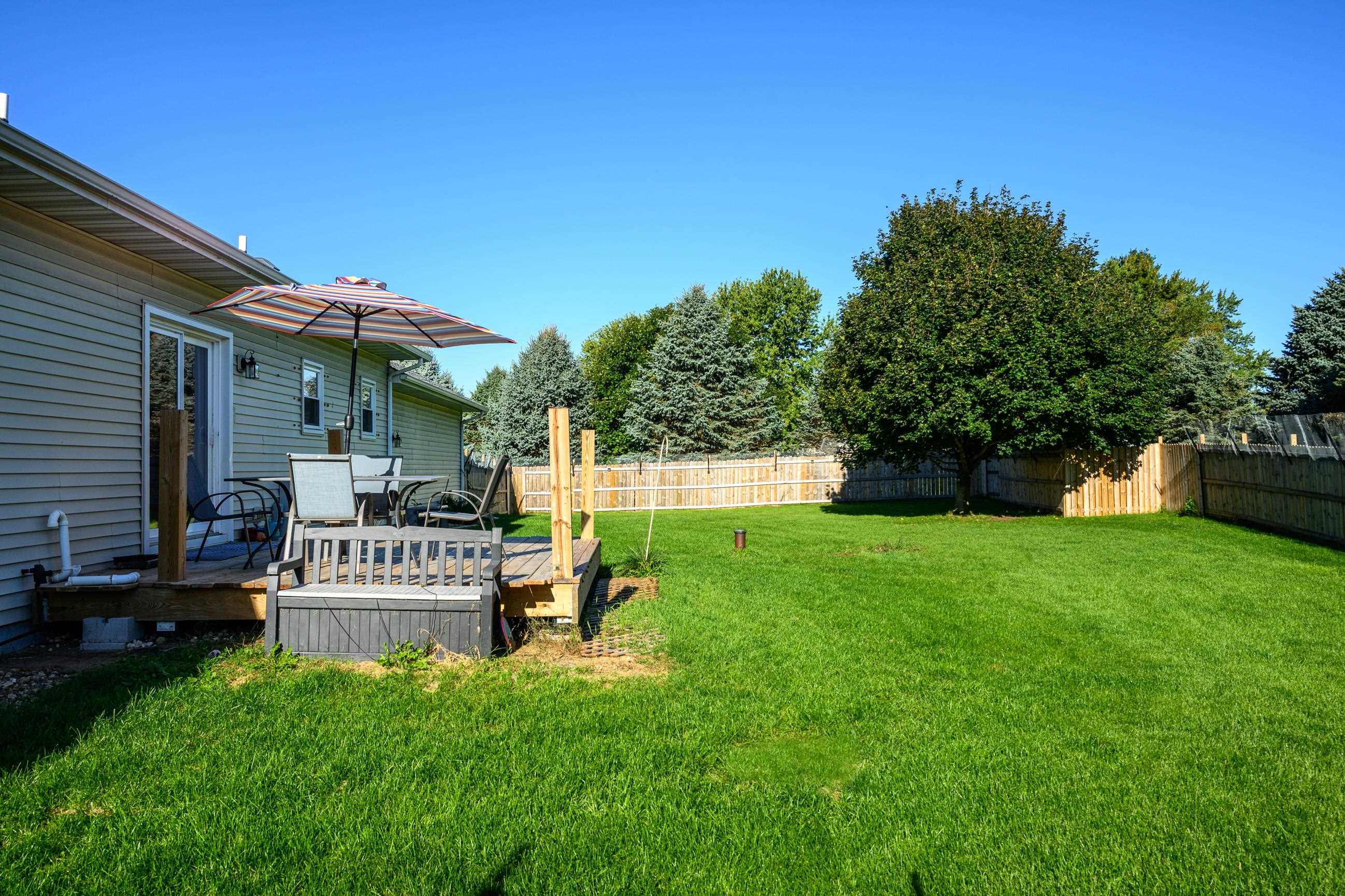 Backyard lawn and fenced yard beside a gray-sided home, with a deck, patio umbrella, and shade tree.
