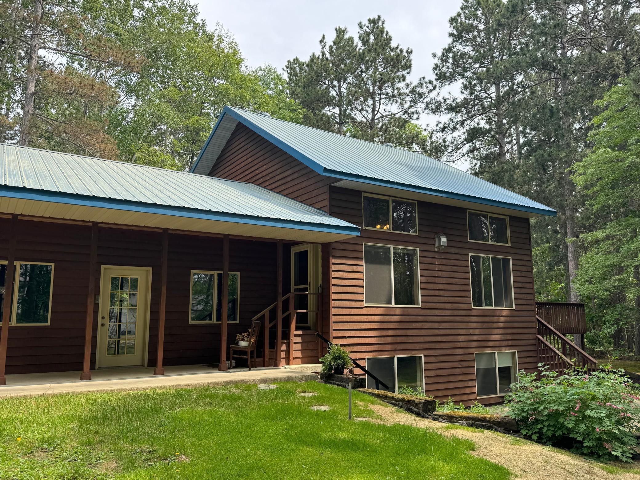 Brown two-story home with blue metal roofs, a covered porch, and mature trees surrounding the lawn.
