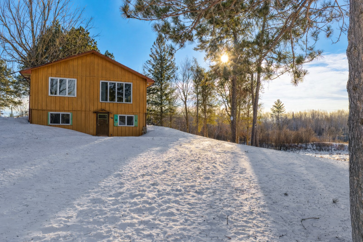 Brown two-story home surrounded by snow and trees in low winter sunlight.