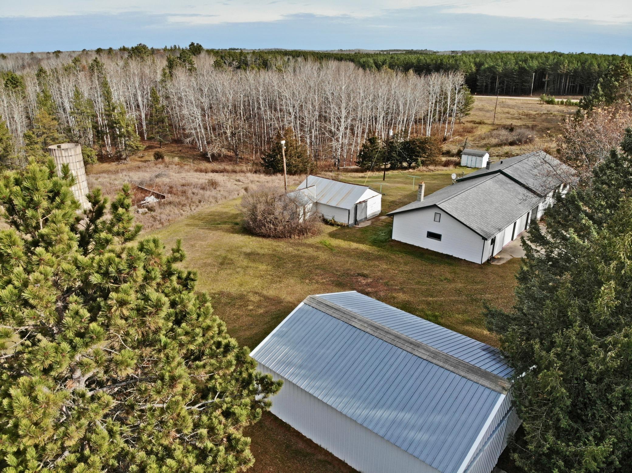 Aerial view of a rural property with a white house, metal-roof outbuildings, a silo, and surrounding woodland.