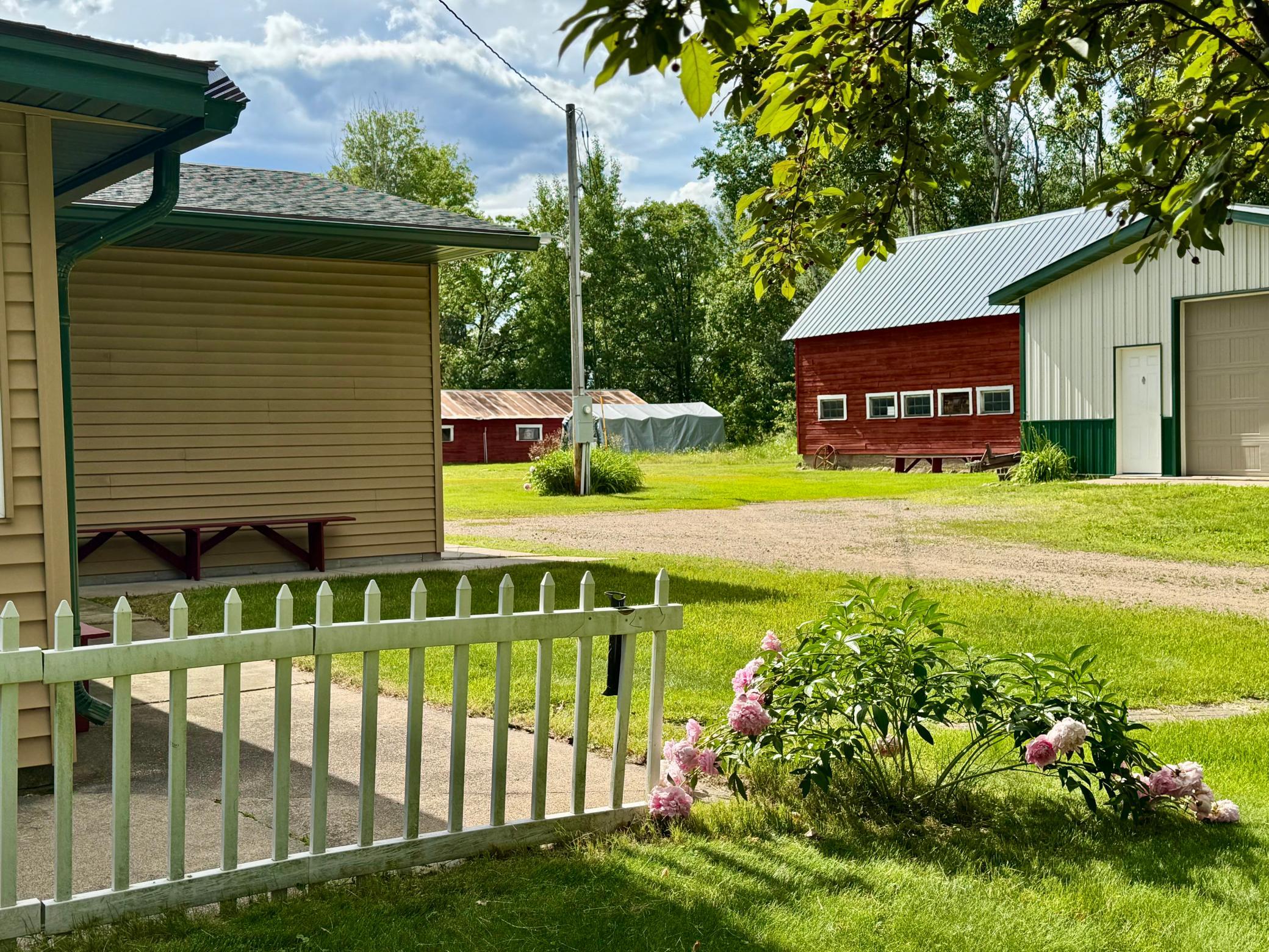 Rural property with a red barn, white garage, picket fence, and pink flowering shrubs.