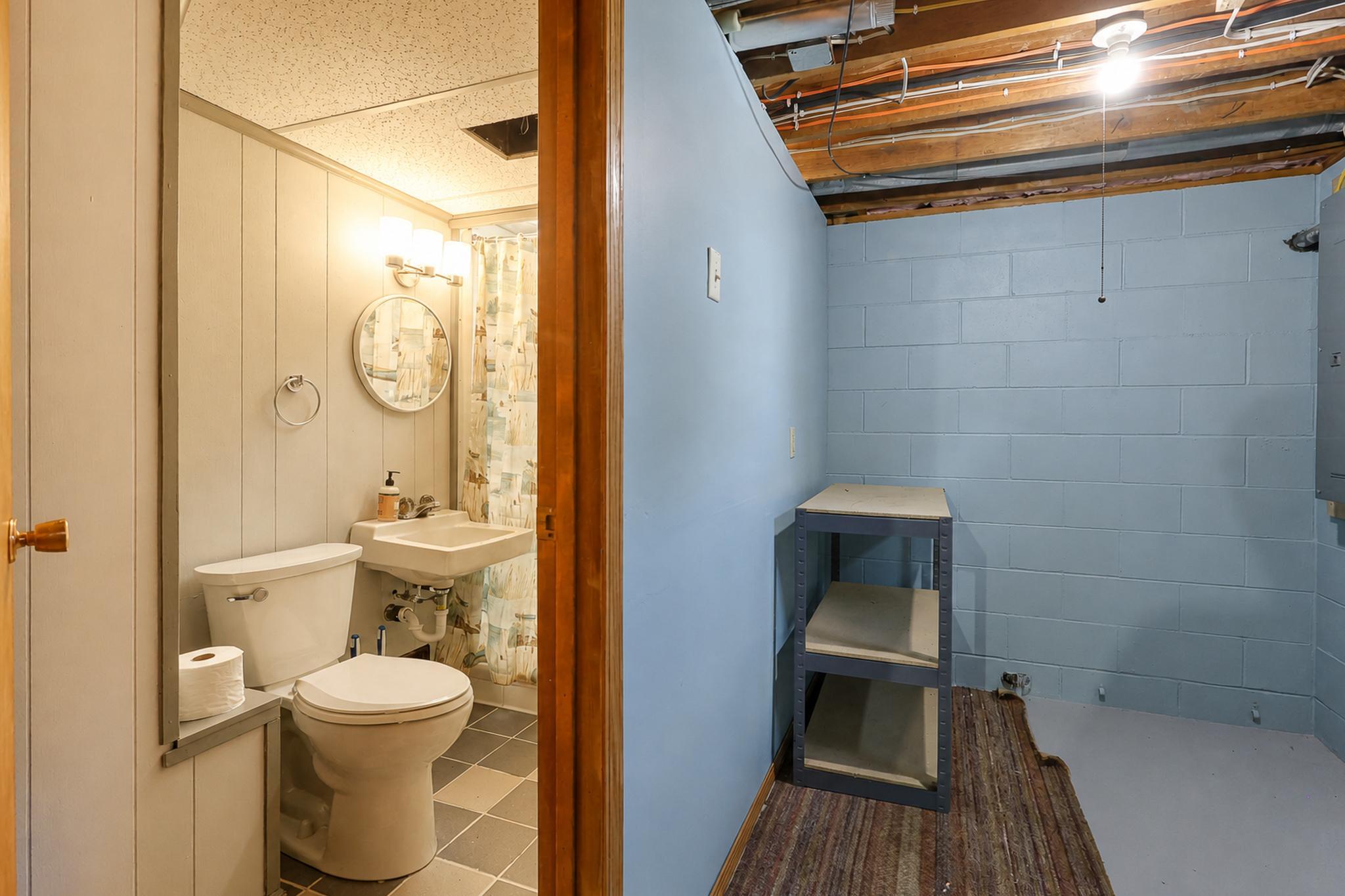 Bathroom with a toilet and sink beside a blue basement room with exposed ceiling joists.