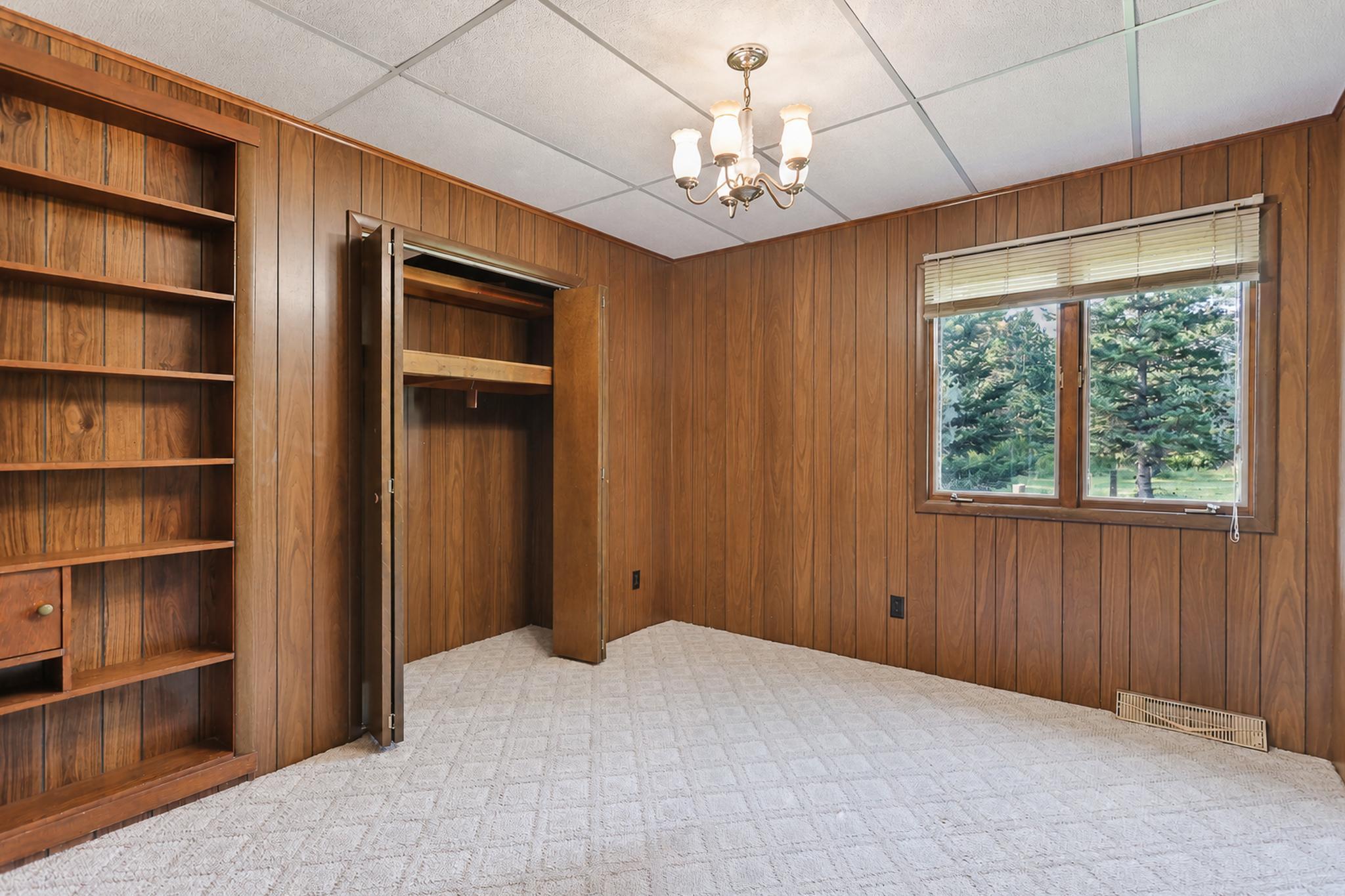 Empty wood-paneled bedroom with built-in shelves, an open closet, and a window overlooking trees.