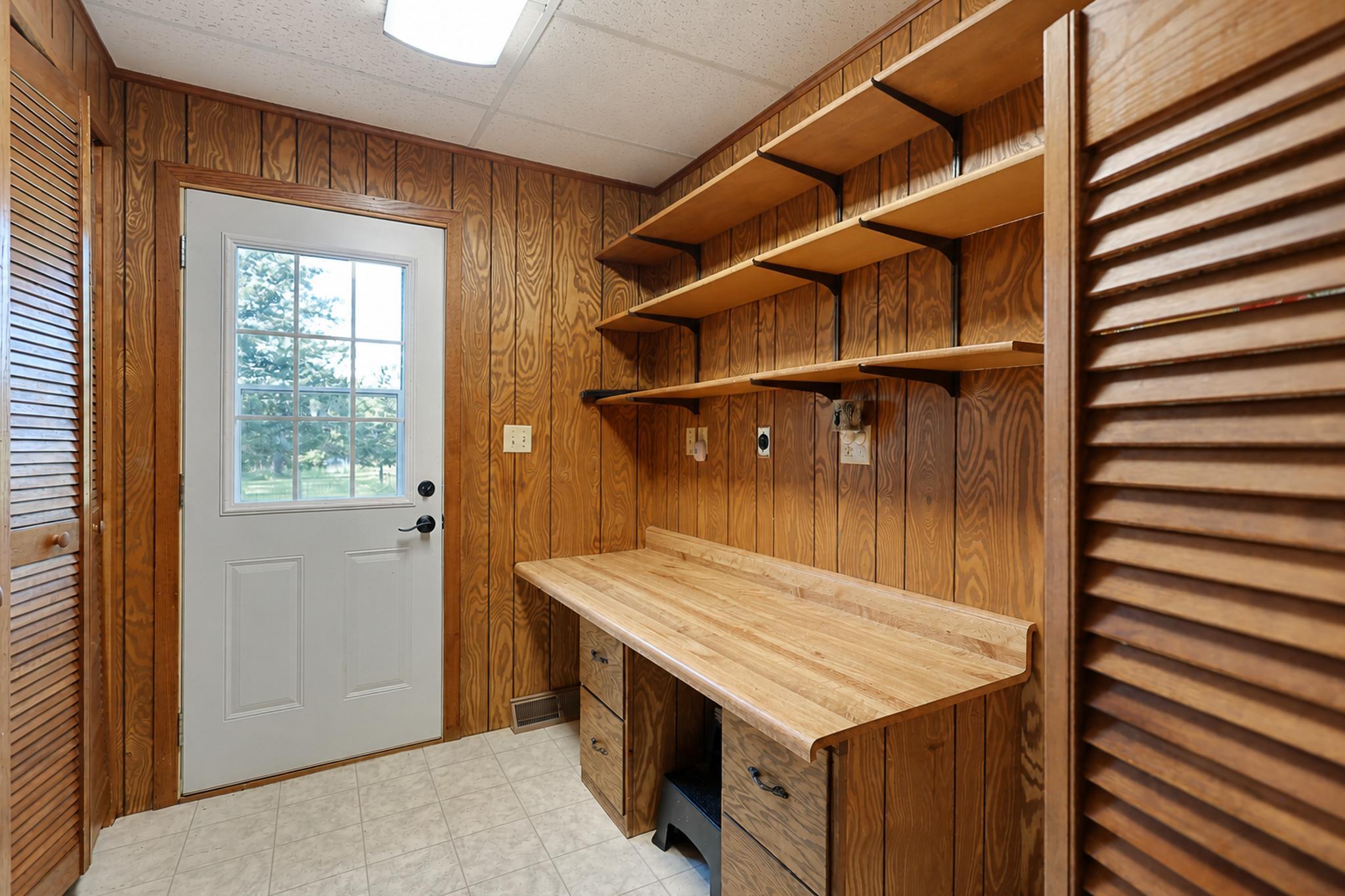 Wood-paneled utility room with built-in work counter, open shelves, and a windowed exterior door.
