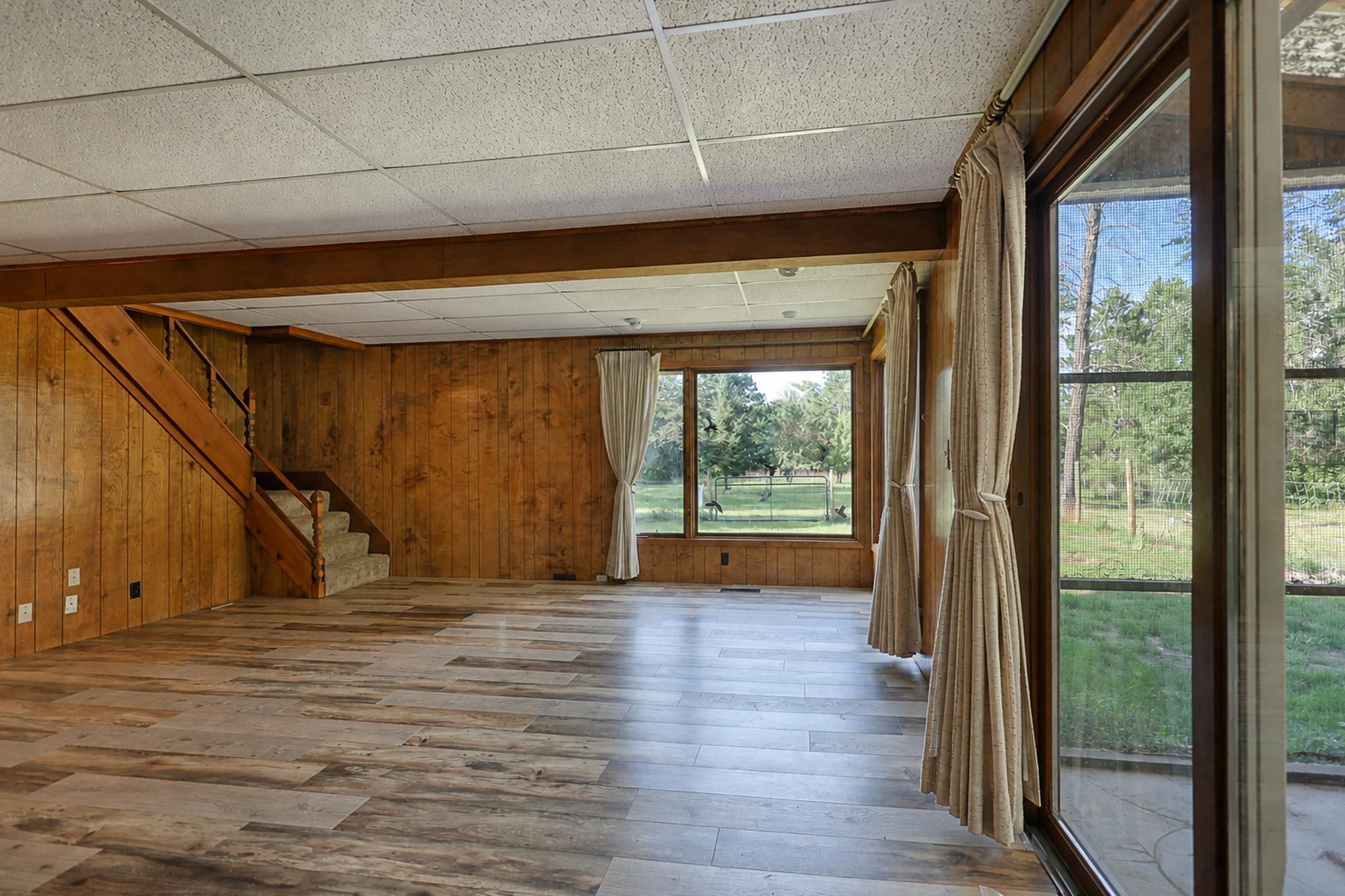 Vacant wood-paneled living room with staircase and large windows overlooking a green yard.