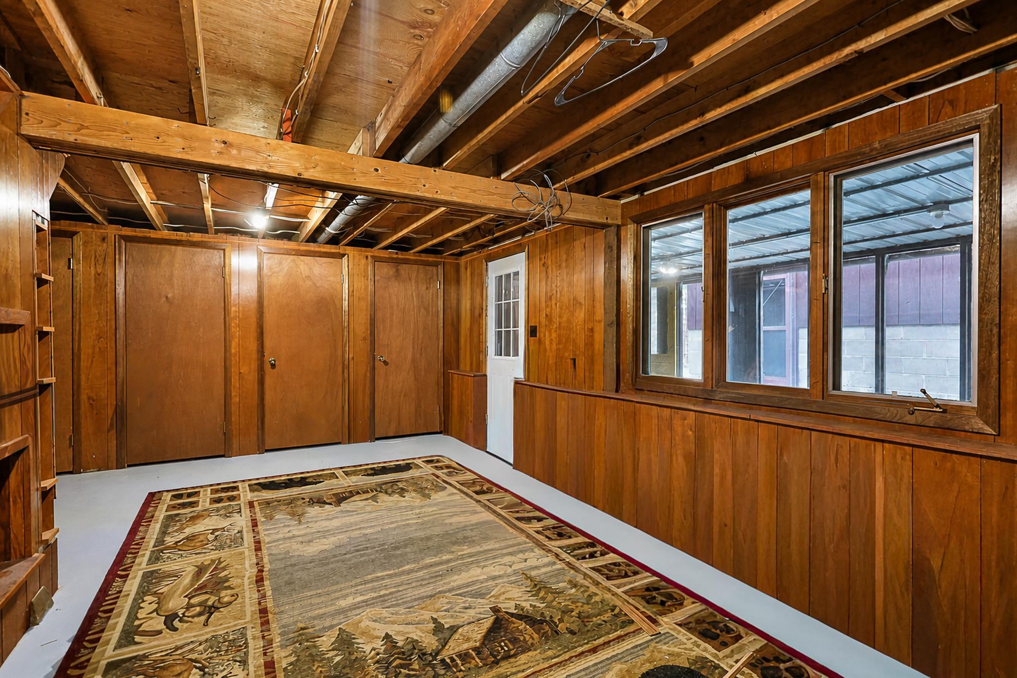 Wood-paneled basement room with exposed ceiling joists, large windows, and a patterned rug.