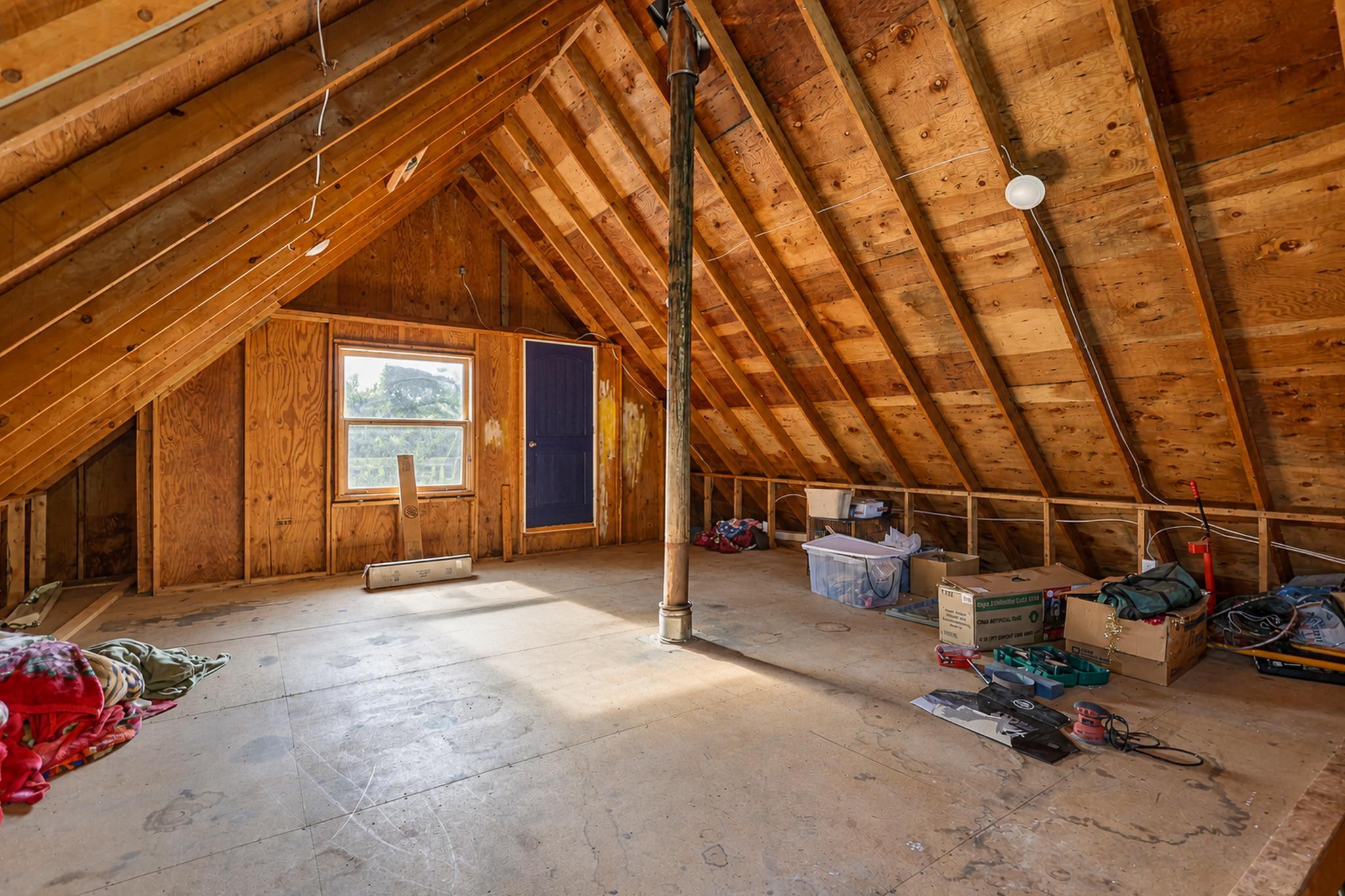 Unfinished attic with exposed wood rafters, a central support post, and boxes and belongings along the edges.
