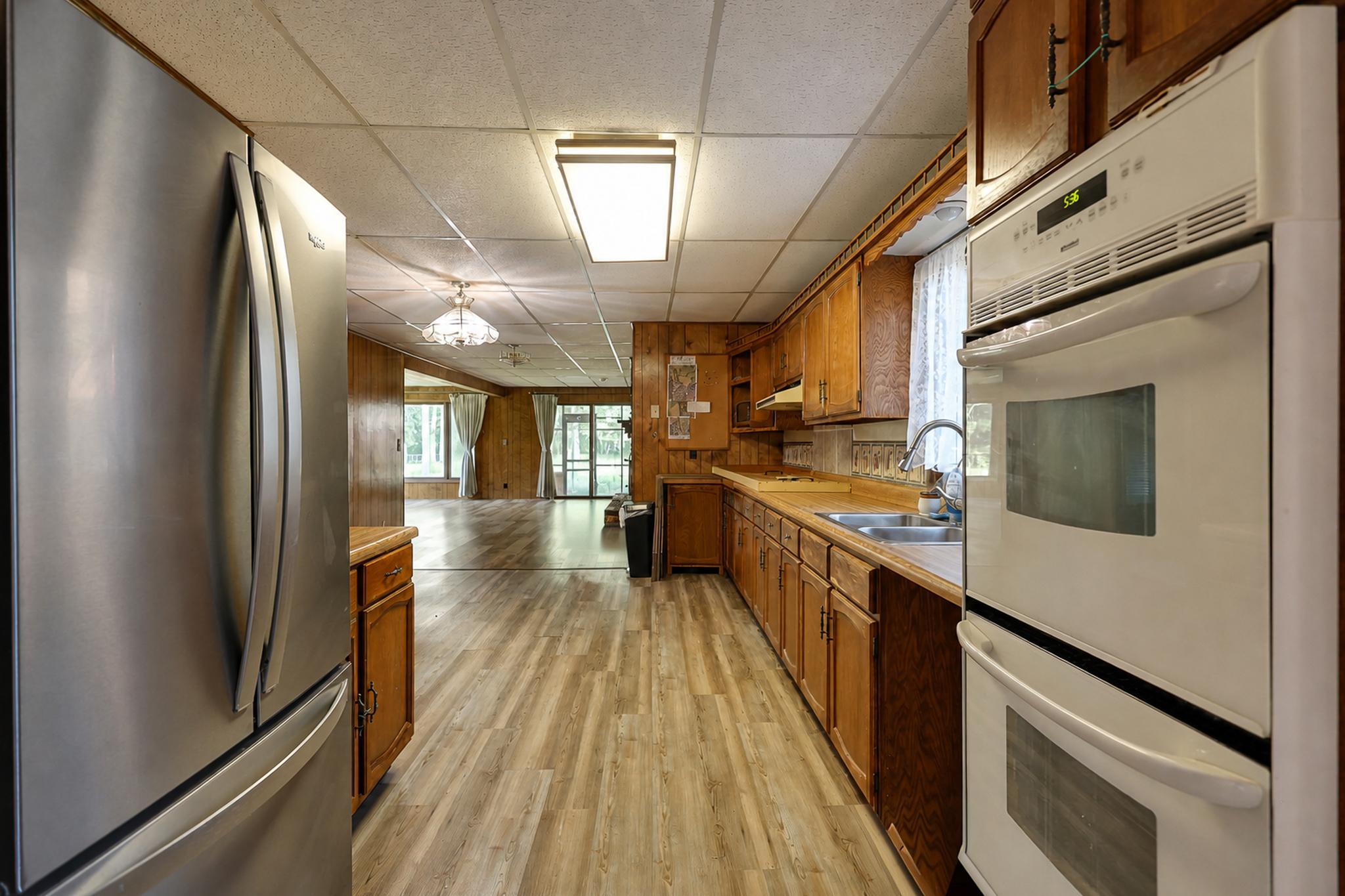 Kitchen with wooden cabinets, stainless refrigerator, and double wall oven opening onto a bright living area.