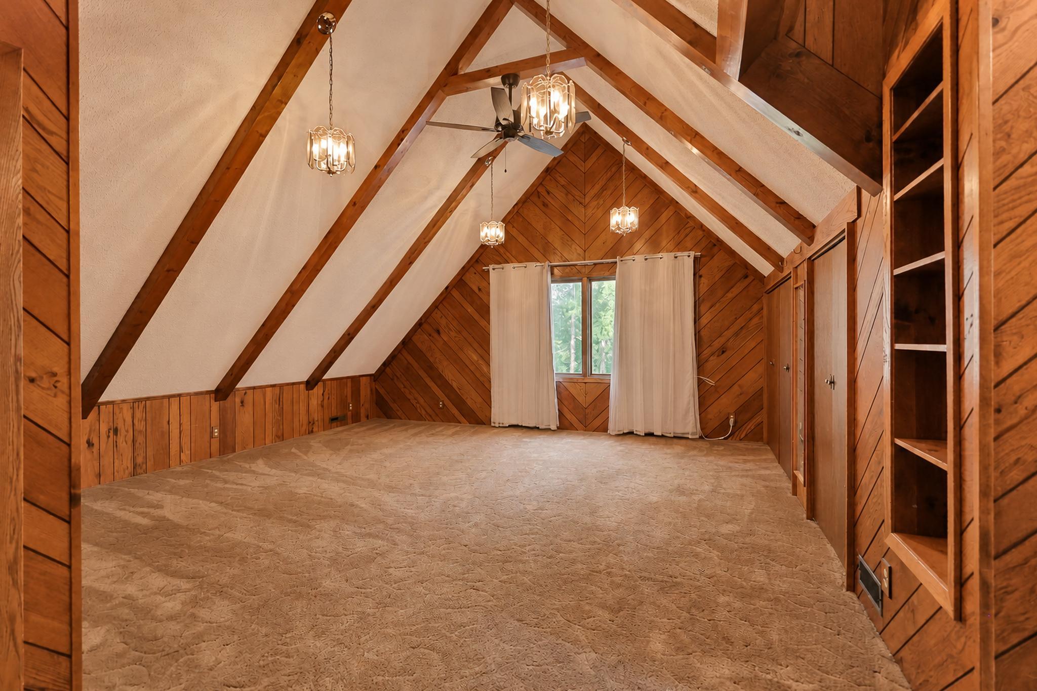 Vacant attic bedroom with exposed wooden beams, warm wood paneling, built-in shelving, and beige carpet.