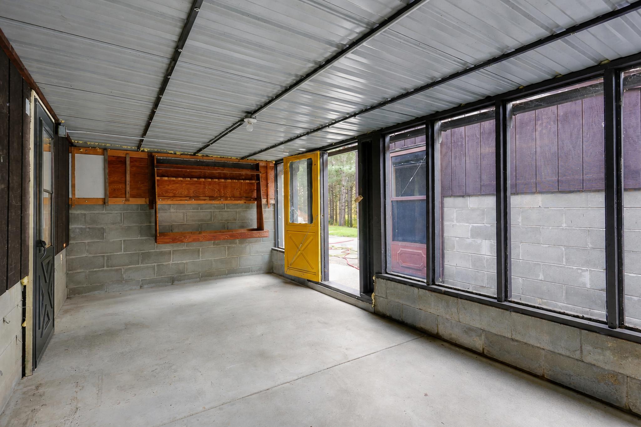 Enclosed porch with a concrete floor, large windows, wooden shelves, and a bright yellow door.