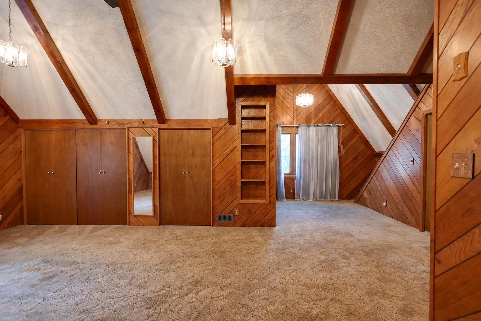 Vacant attic bedroom with wood-paneled walls, exposed beams, built-in closets, and beige carpet.