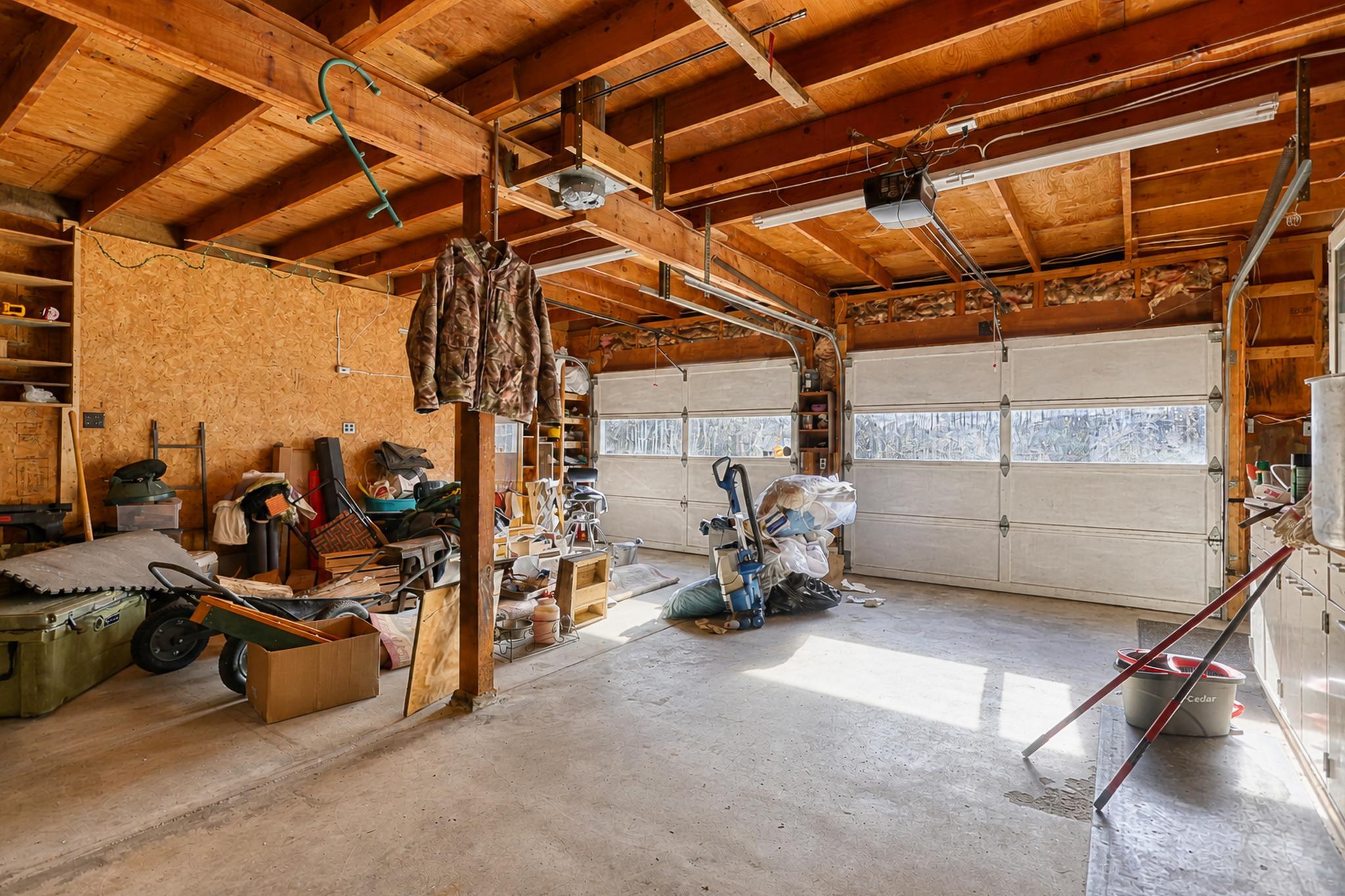 Cluttered garage with exposed timber rafters, two windowed garage doors, and stored equipment.
