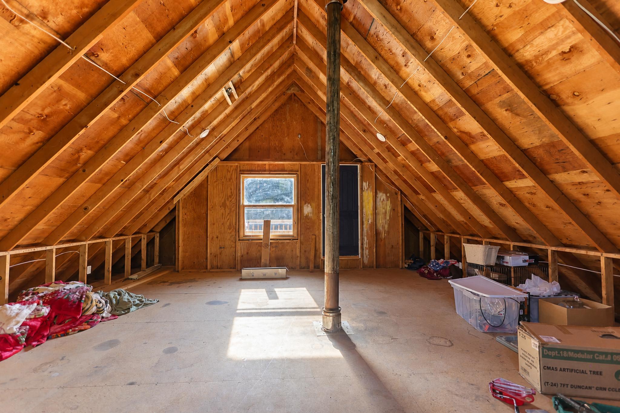 Unfinished attic with exposed wooden rafters, a central stove pipe, and storage items.