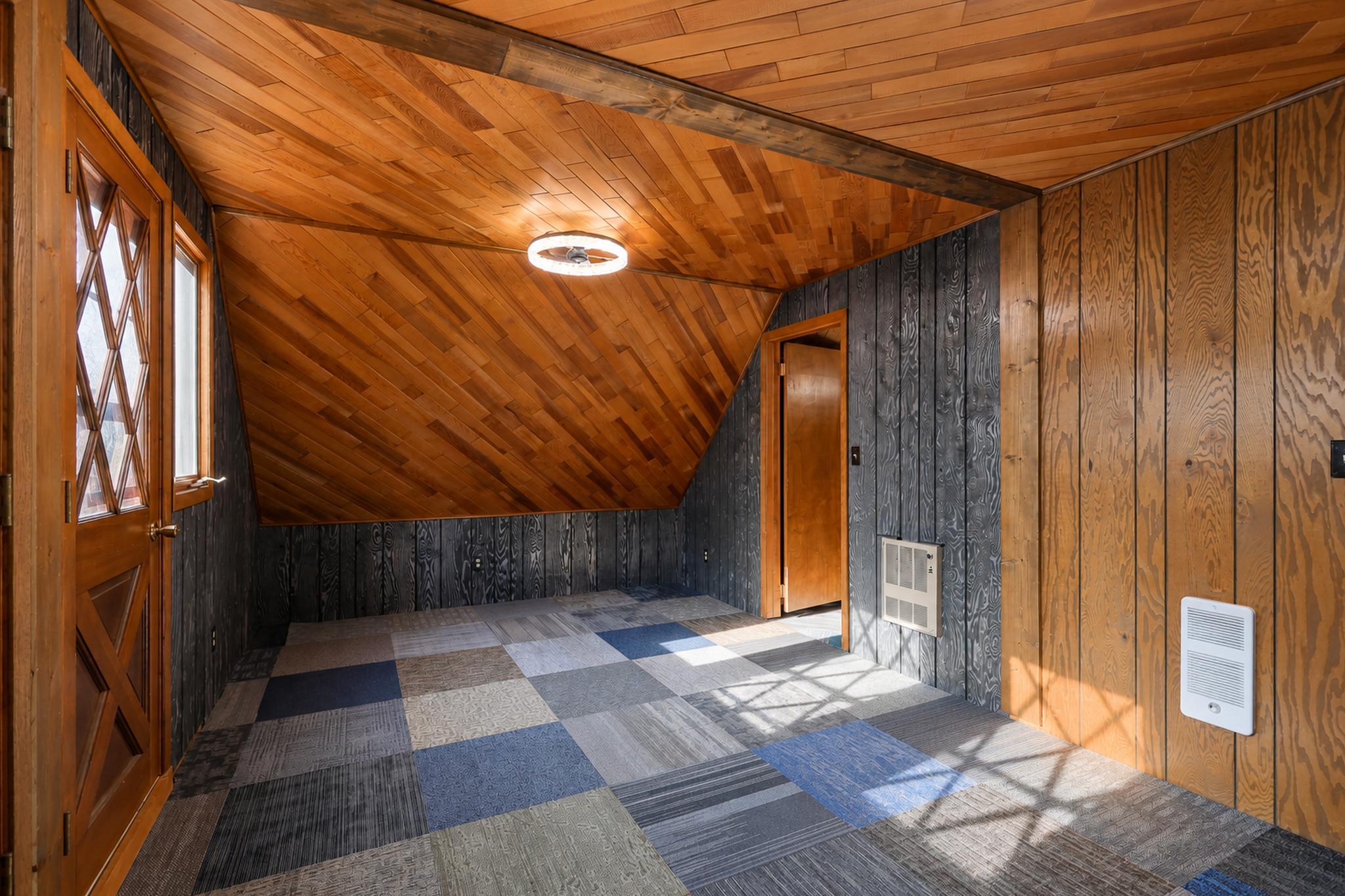Empty room with a sloped wood-paneled ceiling, patchwork carpet, and sunlight through a windowed door.
