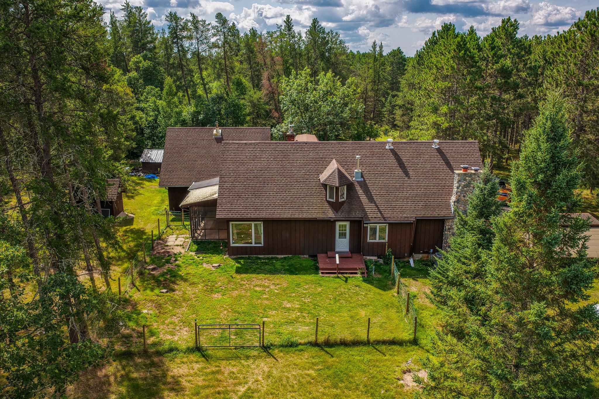 Aerial view of a brown-sided home and fenced grassy yard surrounded by trees.