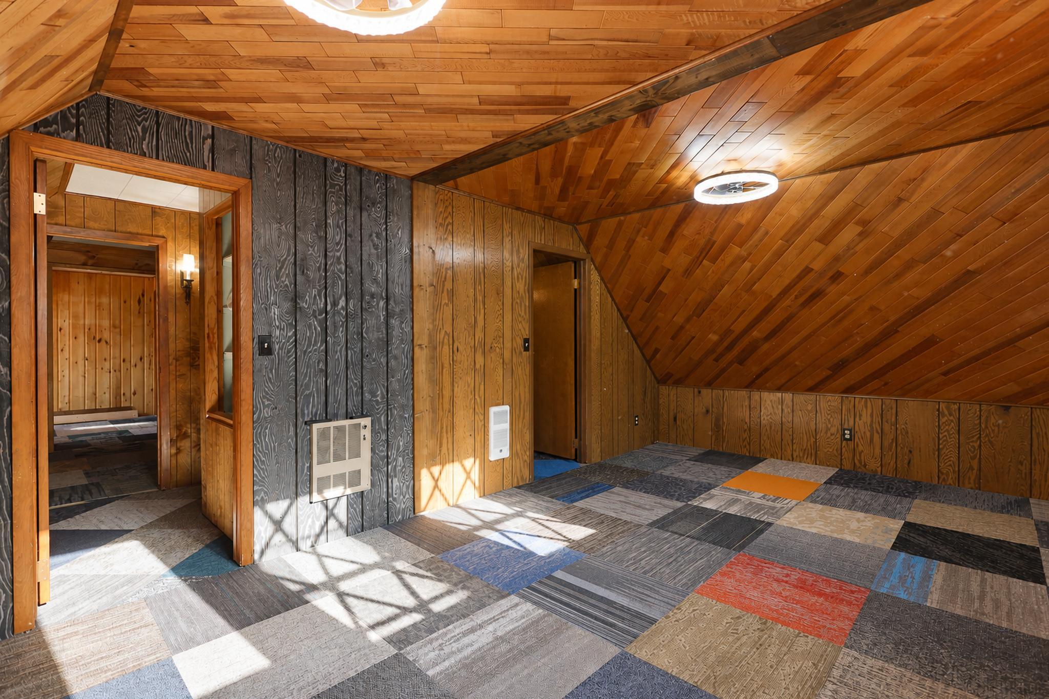 Empty room with a sloped wood-paneled ceiling, multicolored carpet tiles, and doorways to adjoining rooms.