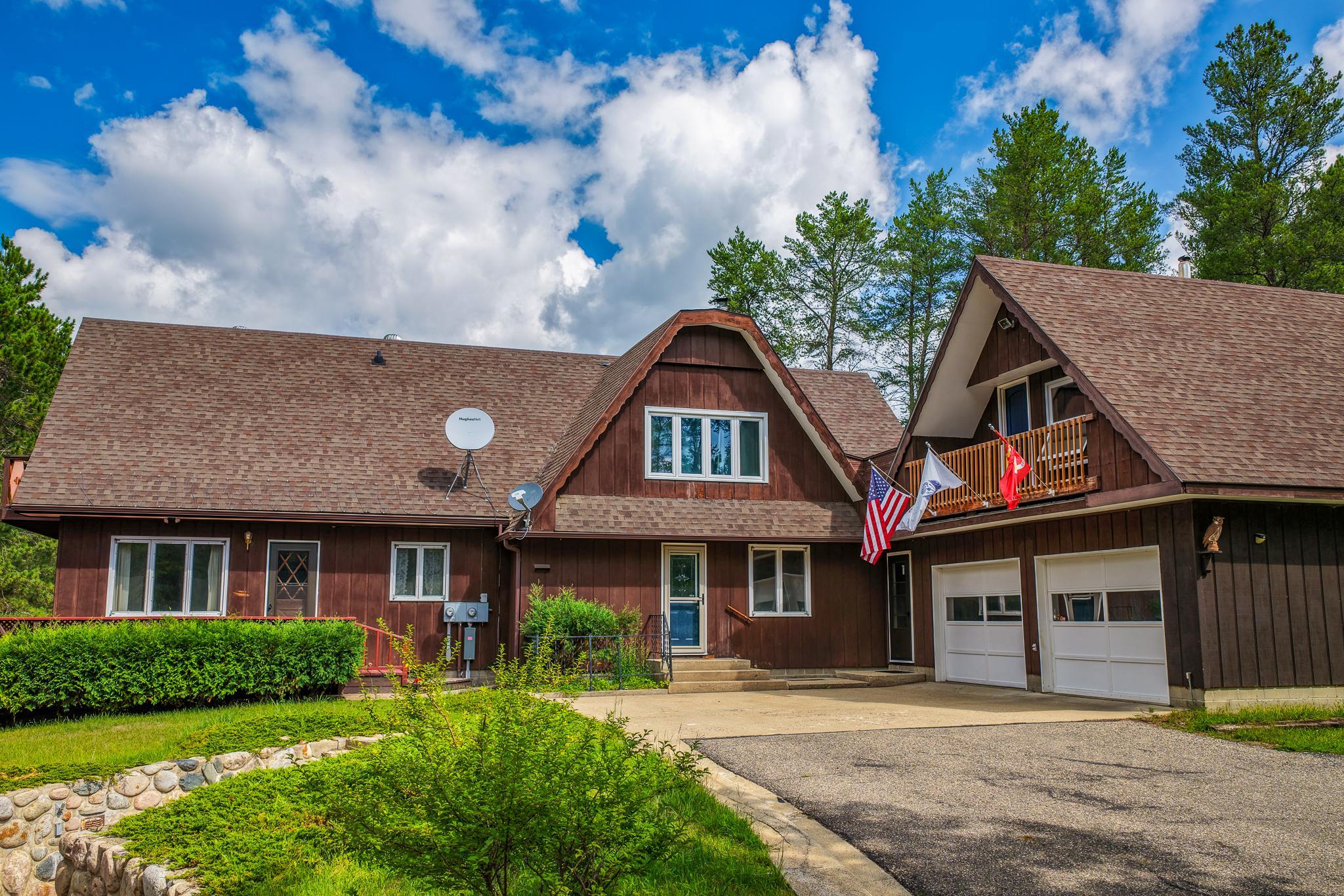 Brown multi-gabled house with a flag-adorned balcony, attached garage, and paved driveway.