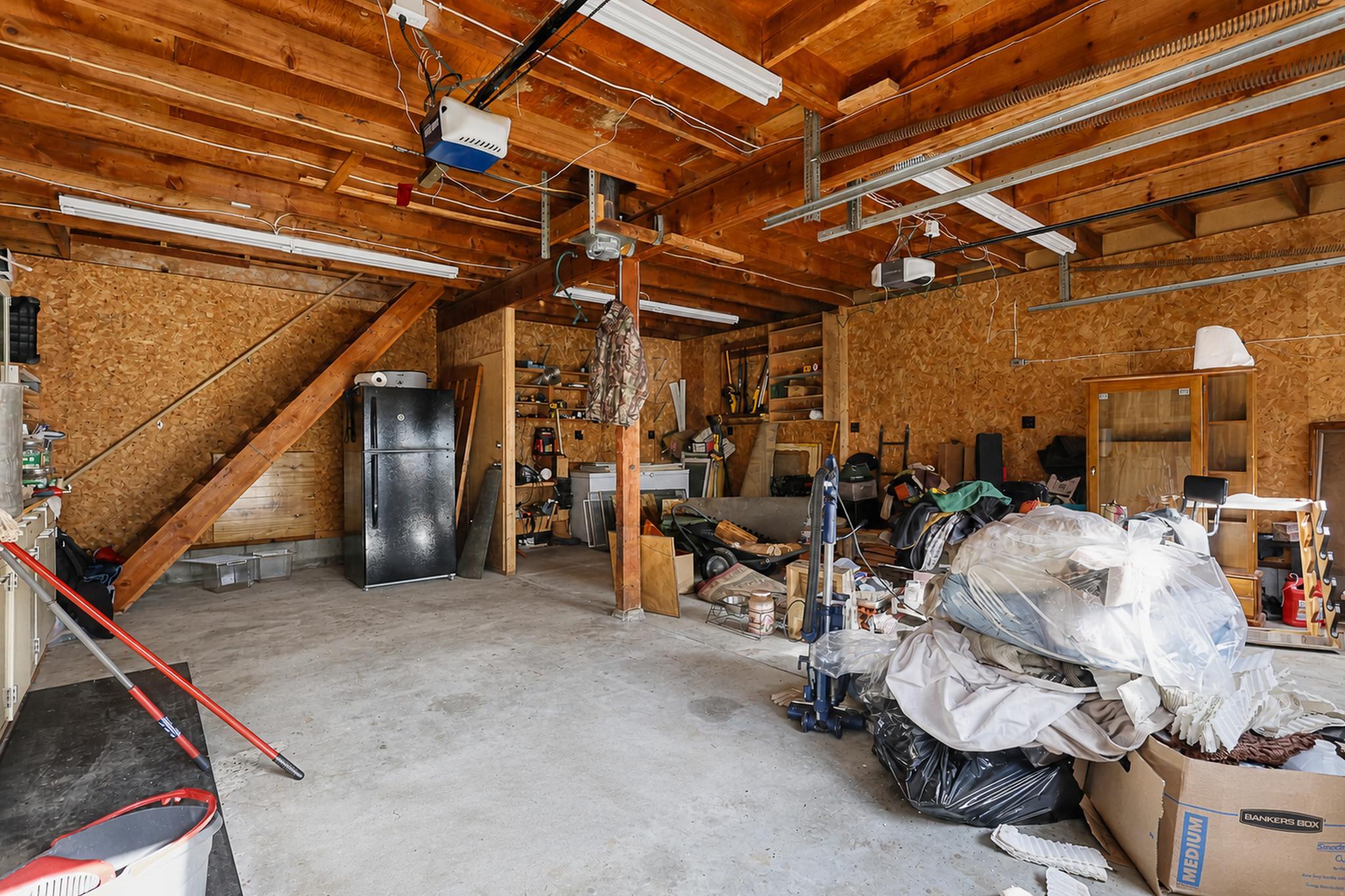 Cluttered garage with exposed wood framing, overhead door tracks, shelving, and stored furniture.