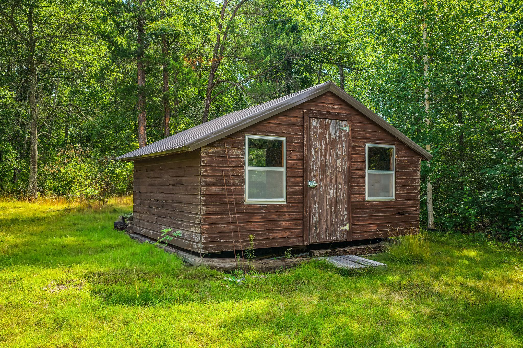 Weathered wooden outbuilding with a metal roof in a tree-lined grassy clearing.