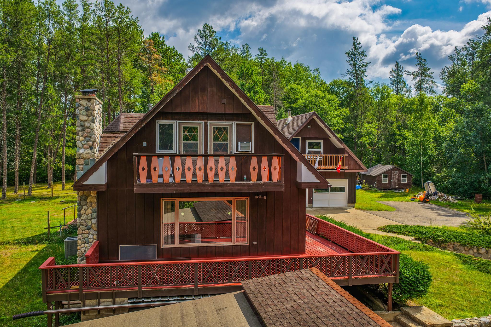 Brown A-frame home with a stone chimney and red-railed decks, surrounded by trees.