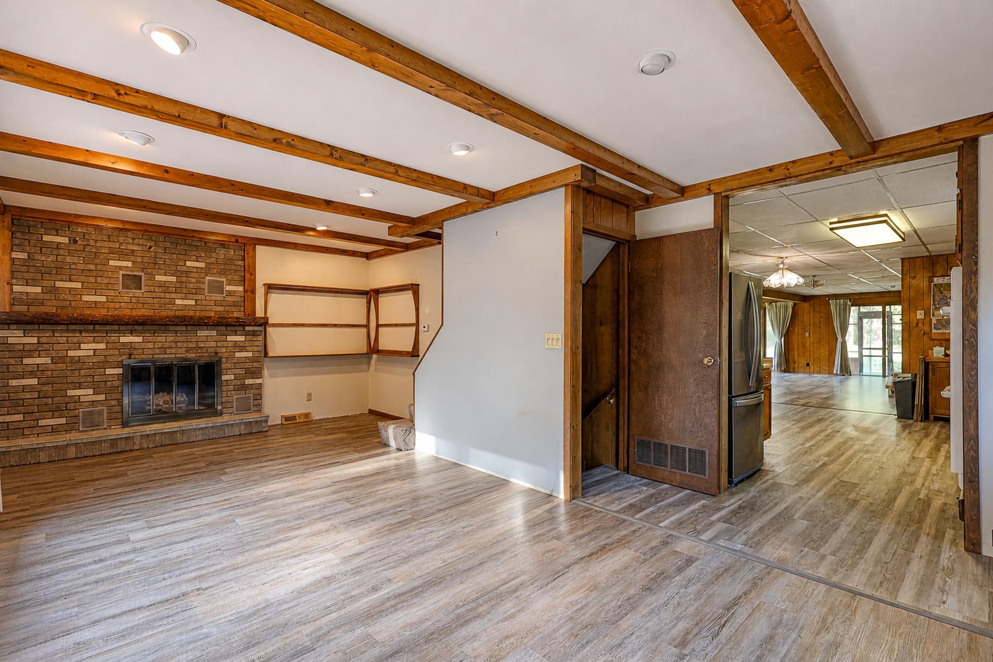 Living room with a brick fireplace, exposed wood beams, and an open passage to a wood-paneled room.
