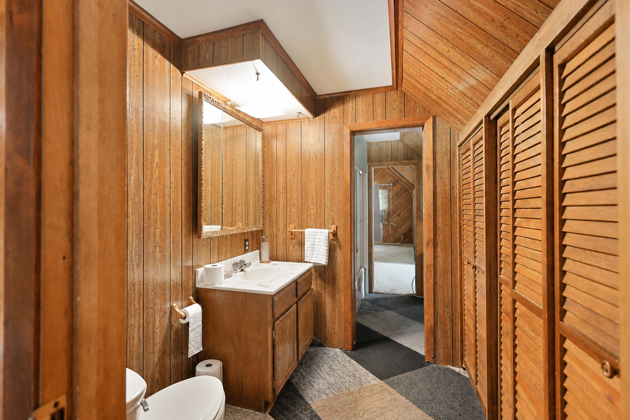 Wood-paneled bathroom with a vanity, louvered closet doors, and an adjoining room visible through the doorway.
