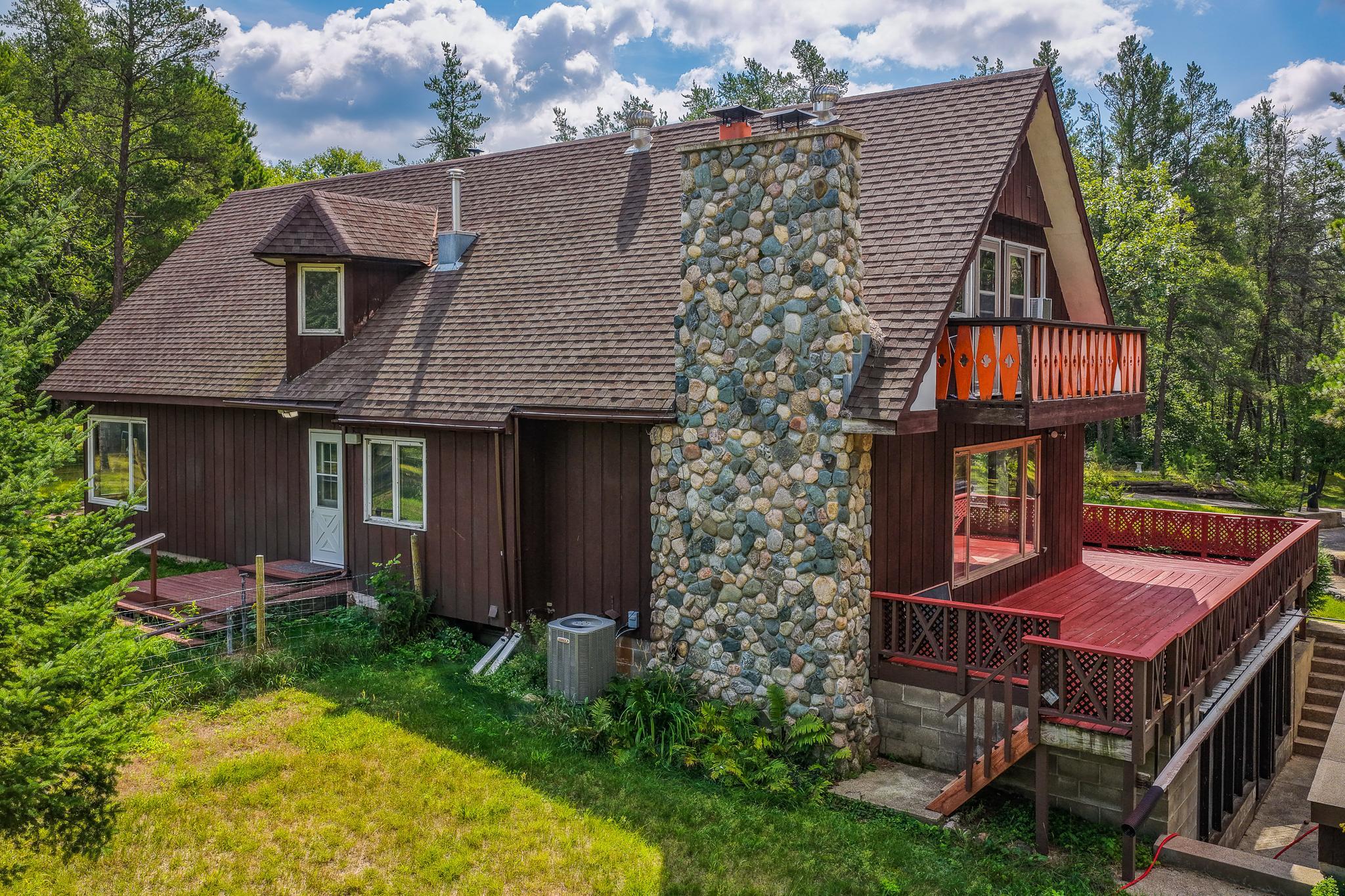 Woodland chalet with a tall stone chimney, orange balcony railings, and a large red deck.