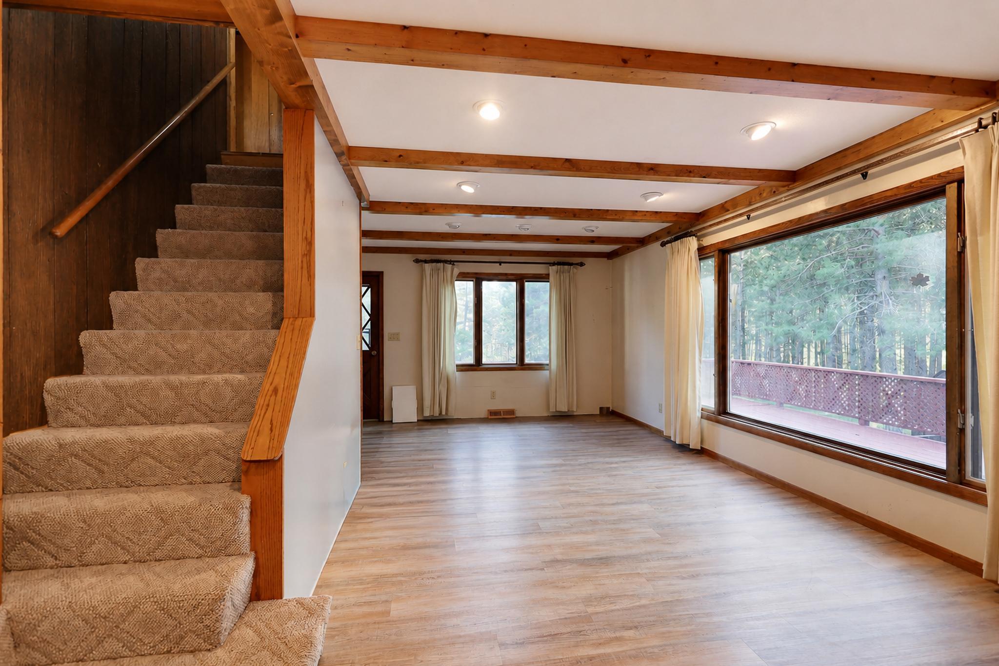 Empty living room with exposed wood beams, carpeted stairs, and large windows overlooking trees.