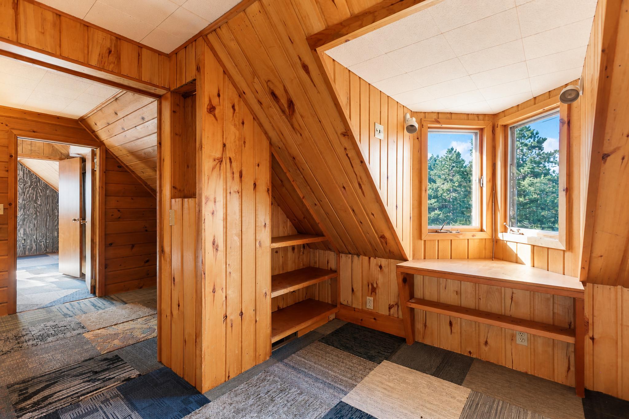 Wood-paneled nook beneath a staircase, with built-in shelves, a window-side desk, and views of evergreen trees.