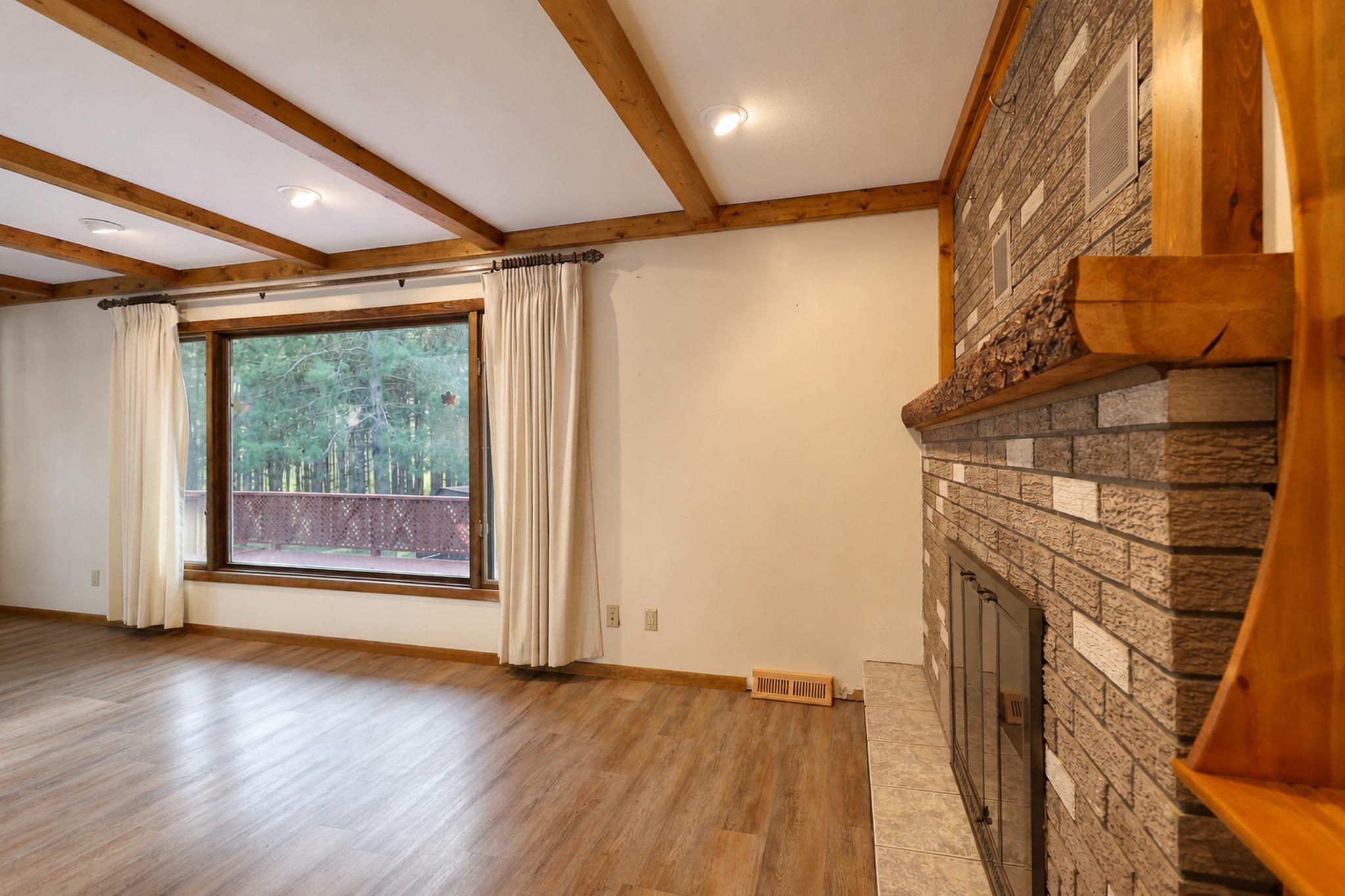 Unfurnished living room with a stone fireplace, exposed ceiling beams, and a large window overlooking trees.