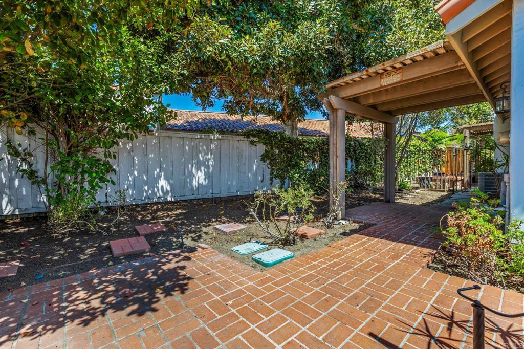 Backyard brick patio beneath a wooden pergola, shaded by leafy trees beside a white fence.