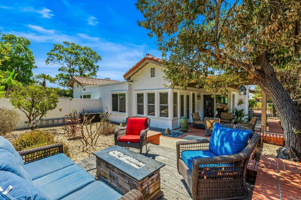 Backyard patio with colorful lounge seating, a stone fire table, and a large shade tree beside the home.