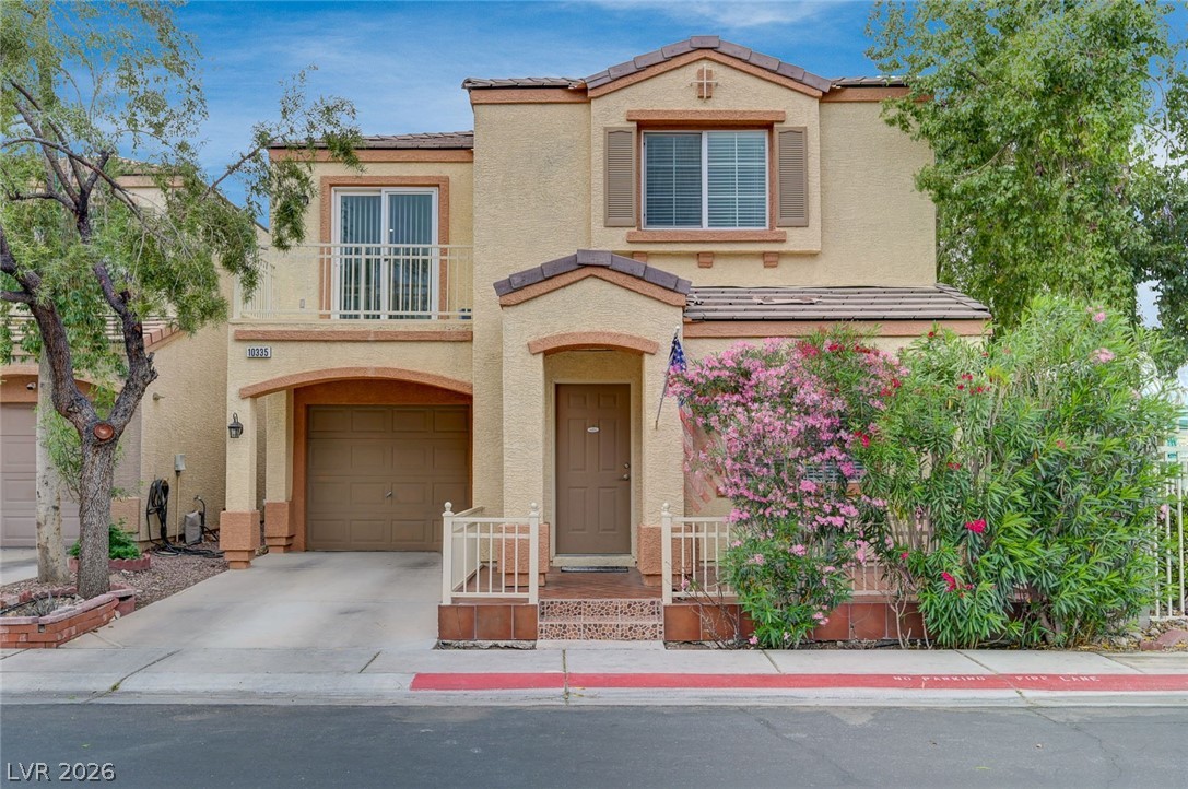 Two-story beige stucco home with a brown garage, small balcony, and pink flowering shrubs.