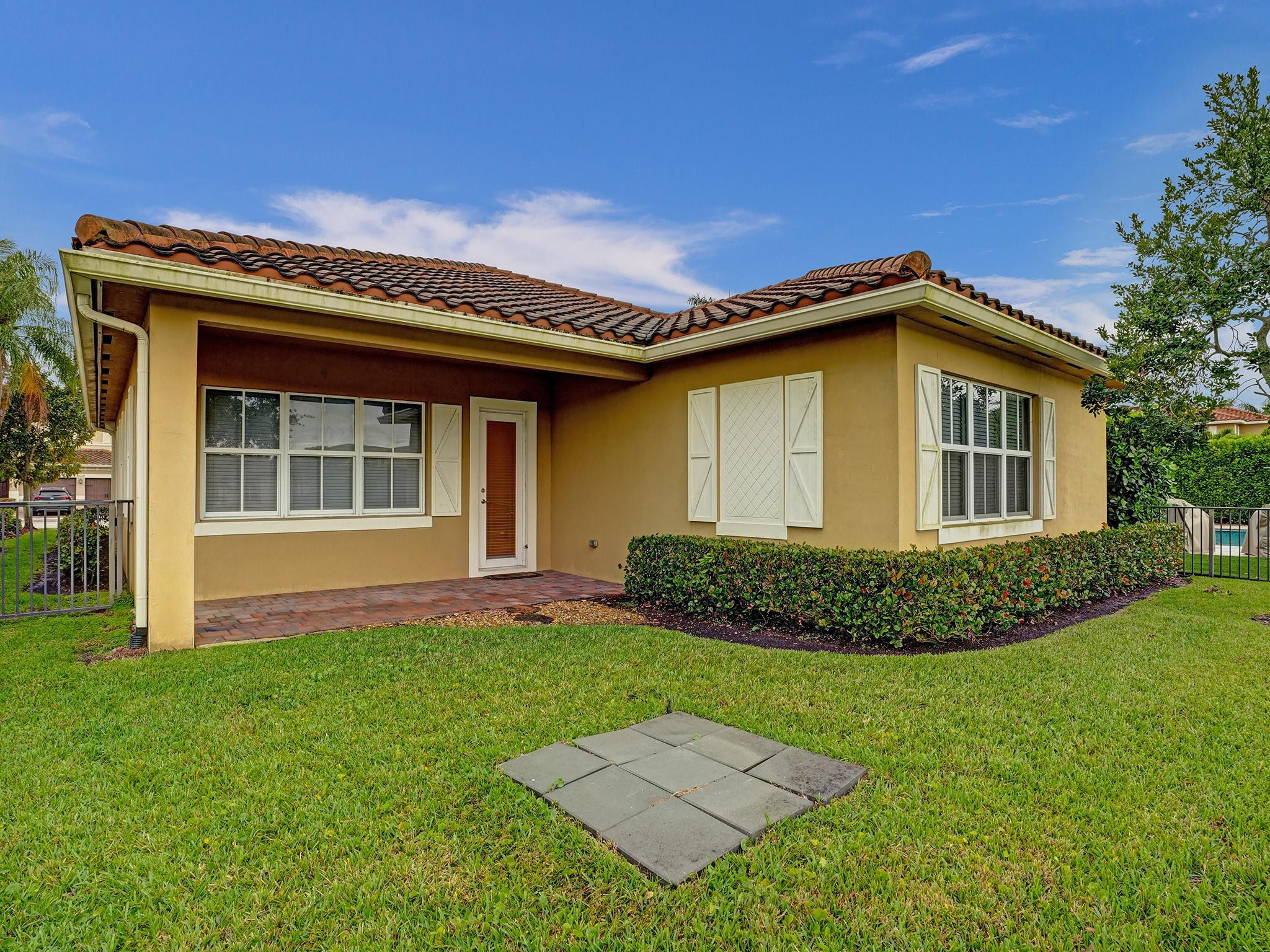 Yellow stucco home with a terracotta tile roof, white shutters, and a hedge-lined lawn beside a pool.