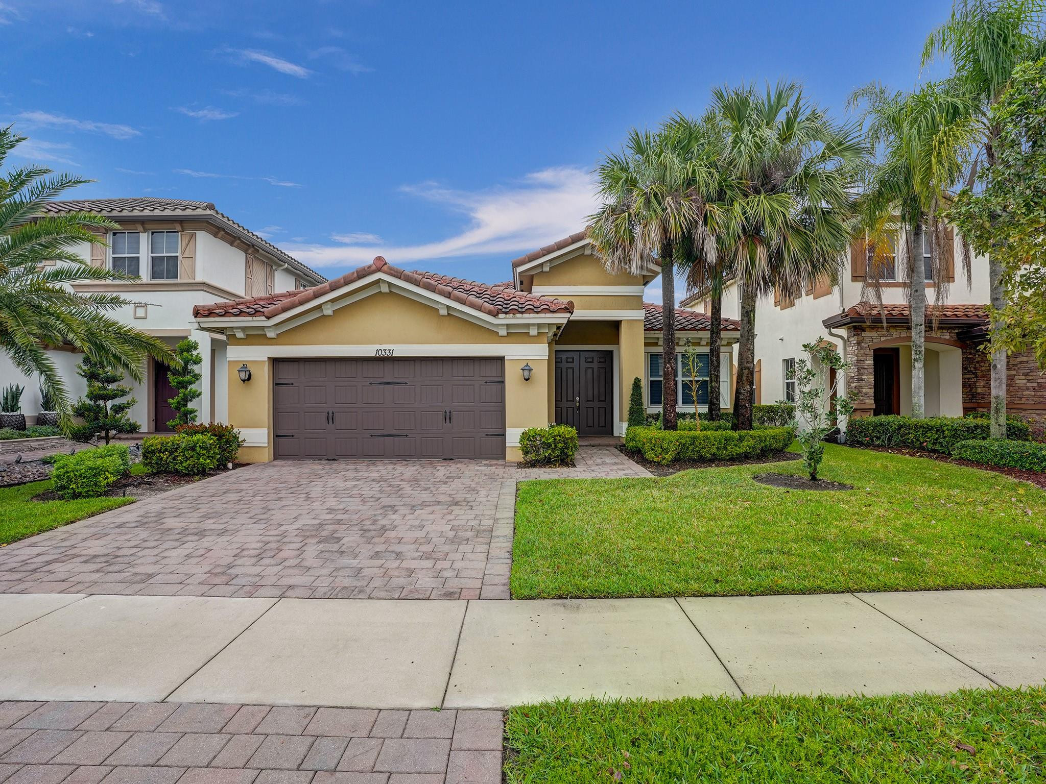 Yellow stucco home with a tiled roof, double garage, paver driveway, and palm trees.
