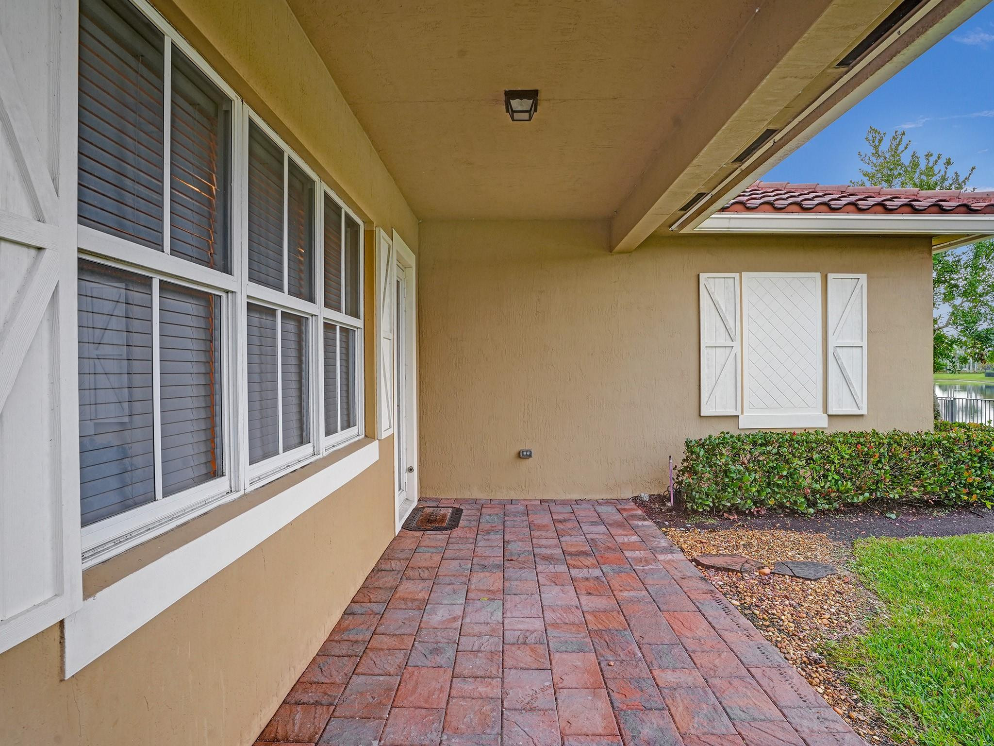 Covered patio with red brick pavers, white-trimmed windows, and a glimpse of water beside the lawn.