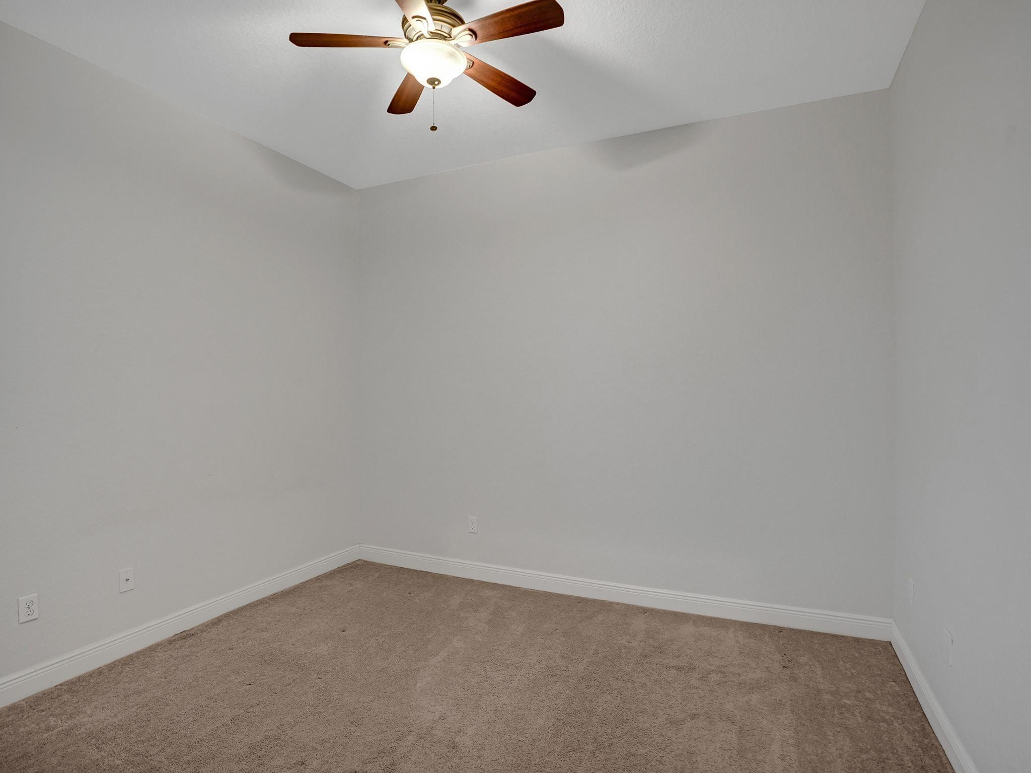 Empty carpeted room with white walls and a lit ceiling fan.